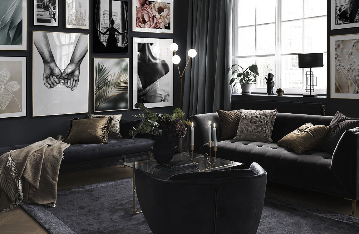 En tavelvägg i ett mörkt vardagsrum med svartvita fotokonstverk, naturmotiv och en rosa pion.