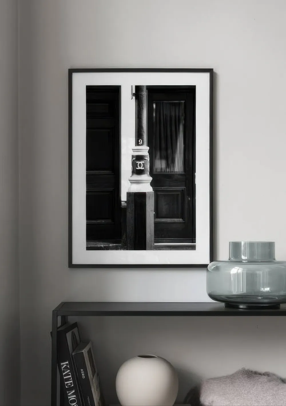 Black and white print of a lamp post with a 9 and a double-C logo, hanging above a black shelf with books and a vase.