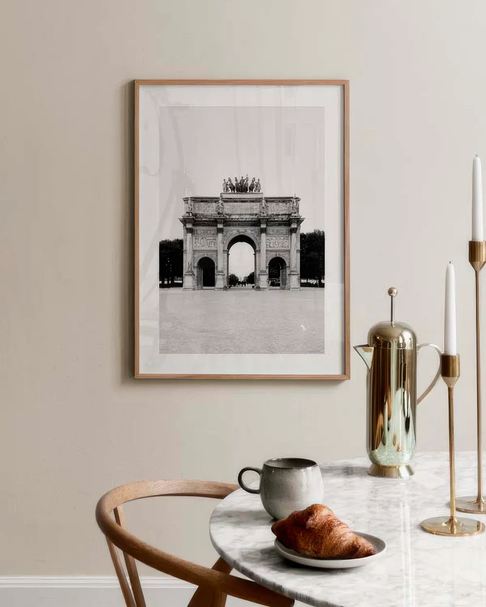 Póster en blanco y negro del Arco del Triunfo de París en una pared beige, con una mesa de mármol, café y un cruasán.