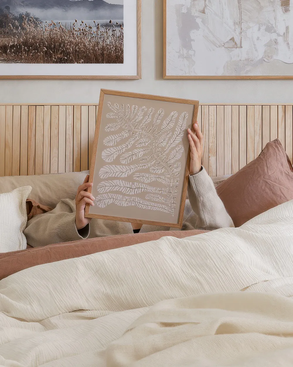 Póster botánico de hojas de helecho blancas sobre fondo beige, sostenido en una cama acogedora con ropa de cama crema y rosa.