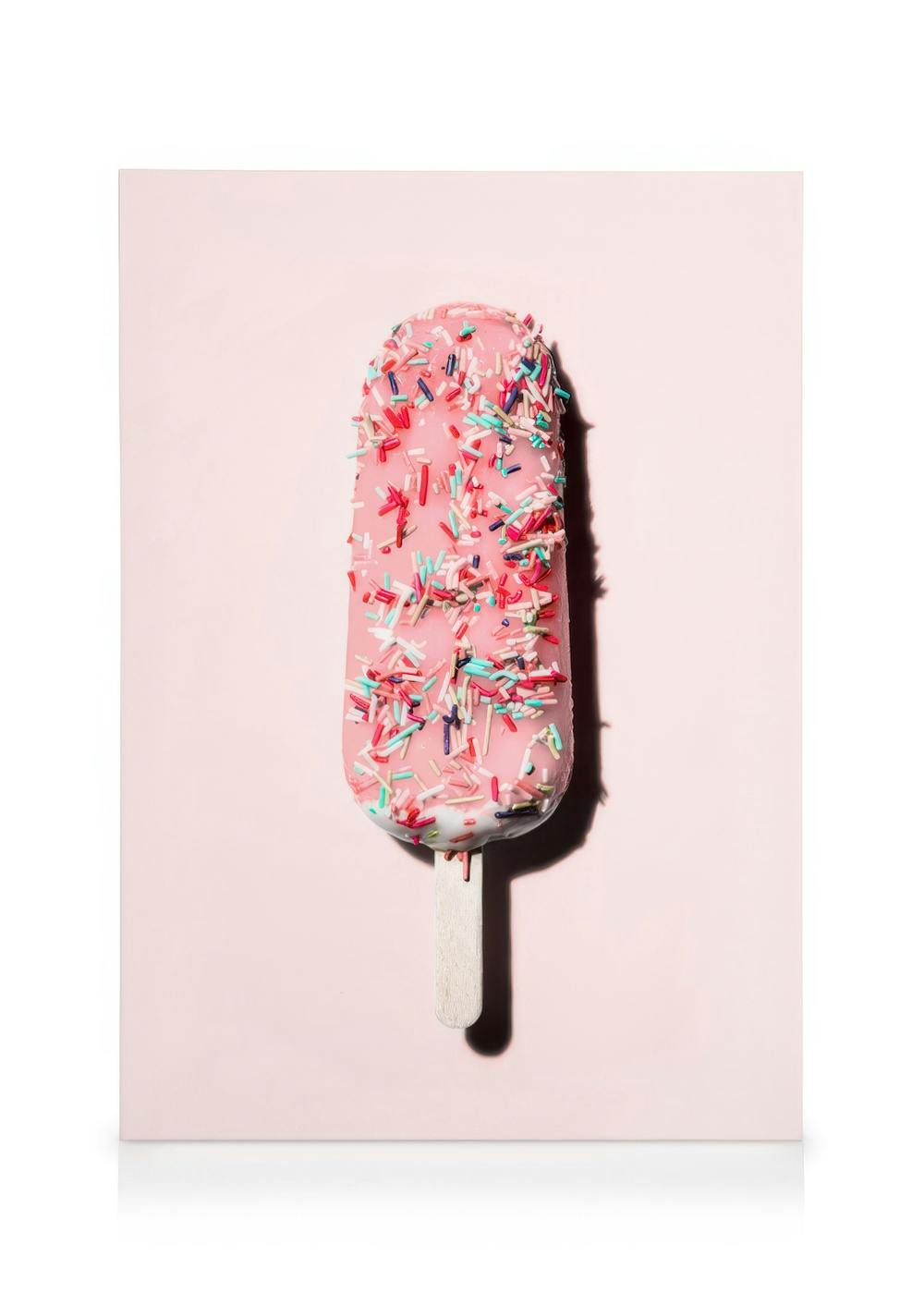 Rosa Eis am Stiel mit bunten Streuseln auf hellrosa Hintergrund, Pop-Art-Stil.