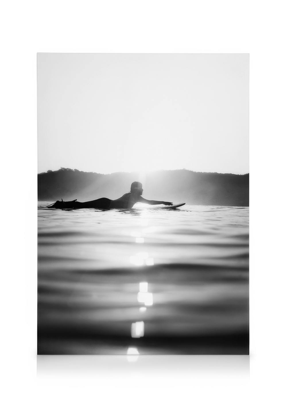Stampa tela in bianco e nero di surfista che galleggia sullacqua, con riflessi di luce e colline sullo sfondo.