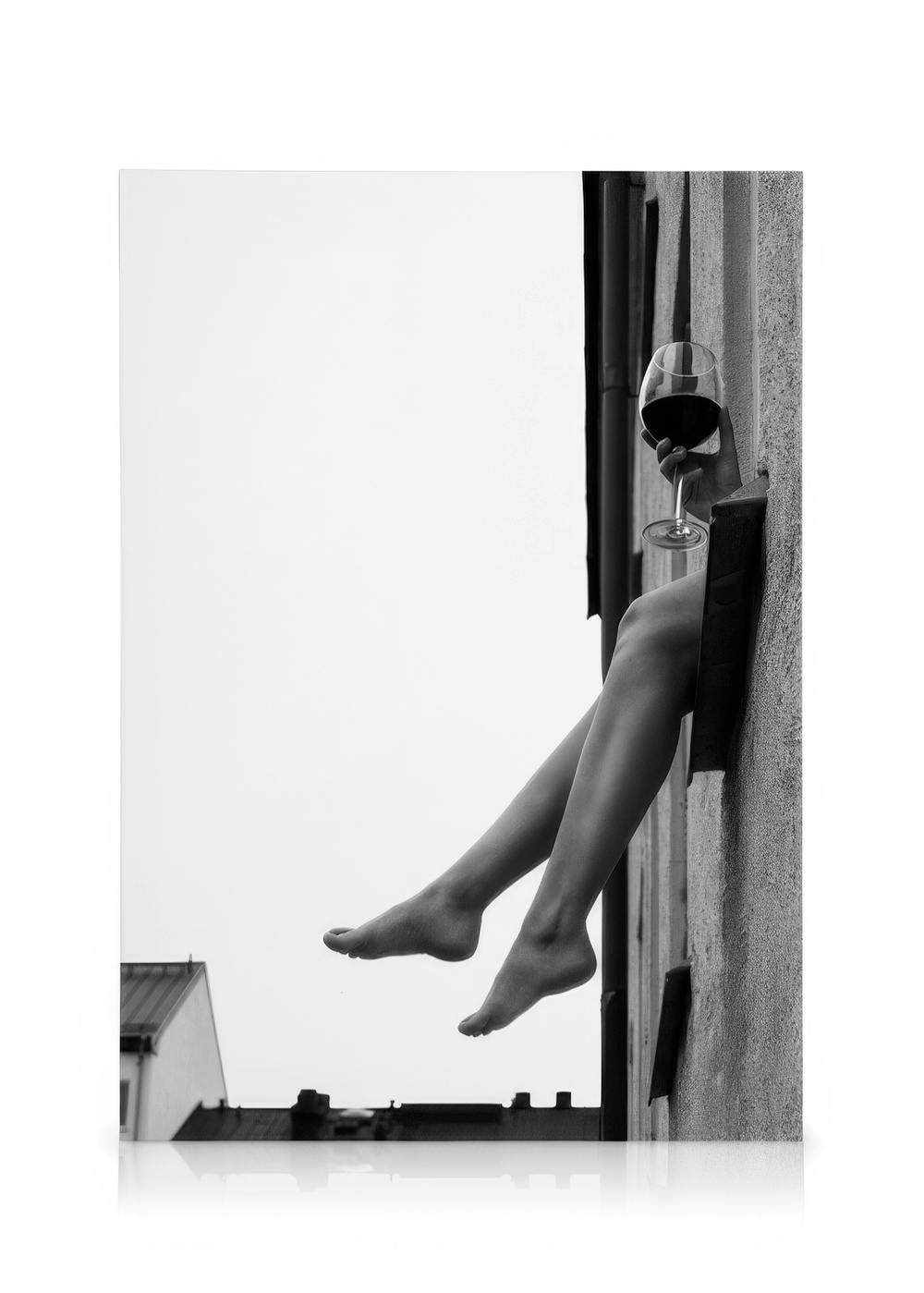 Schwarz-Weiß-Leinwandbild. Eine Person genießt Wein, Beine und Glas ragen aus einem Fenster, Dächer im Hintergrund. Wein im Himm