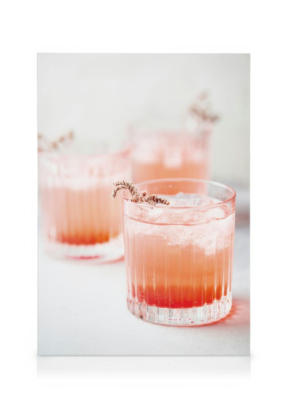 Pink lemonade drink in a fluted glass with ice and a dried lavender garnish, two more blurred glasses in the background