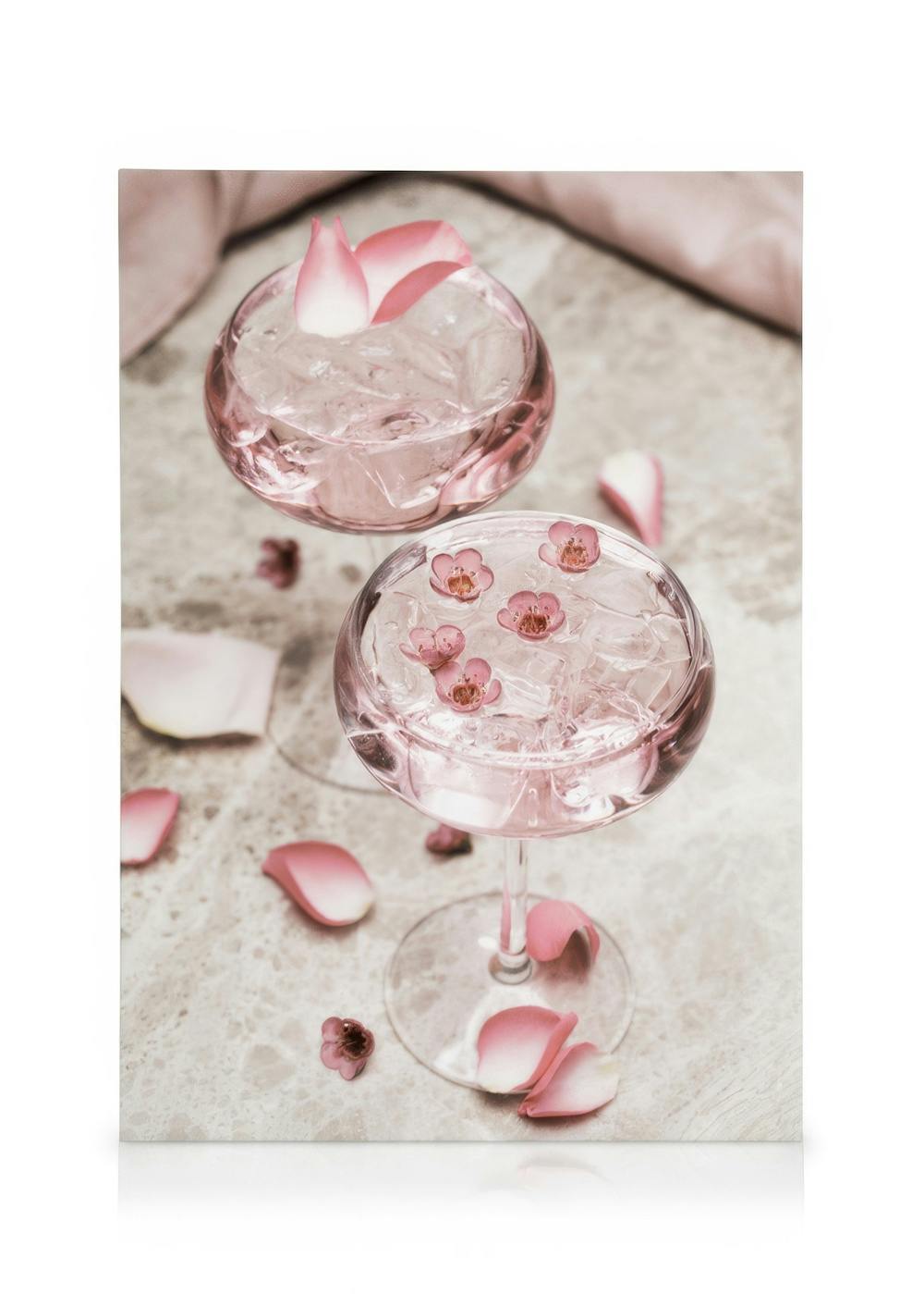 Zwei rosa Cocktails mit Eis und Blütenblättern in eleganten Gläsern auf hellem Marmorhintergrund mit Rosenblättern.