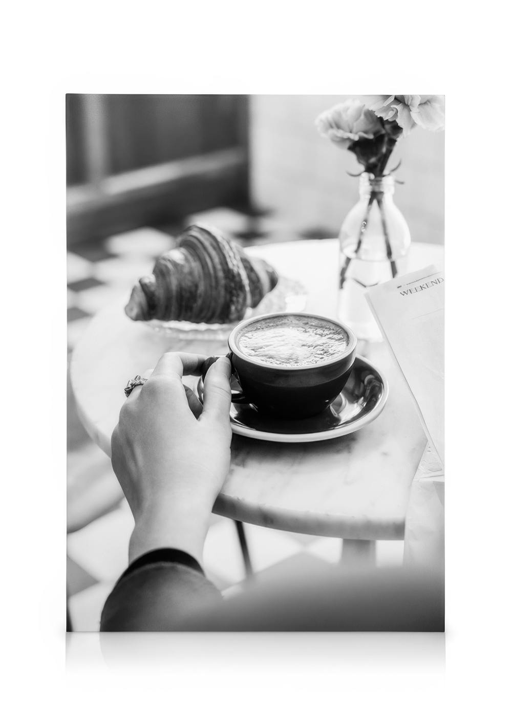 Μαύρο-άσπρο καμβάς με χέρι που κρατά φλιτζάνι καφέ, κρουασάν και λουλούδια σε τραπέζι