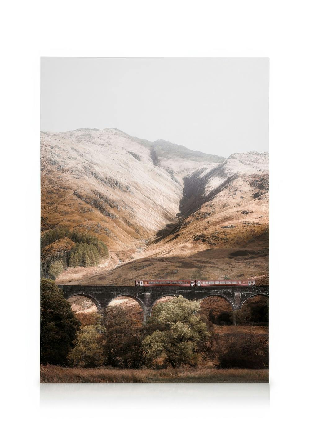 Red train on stone bridge, snow-covered mountains, green trees, light sky. Highland train canvas print.