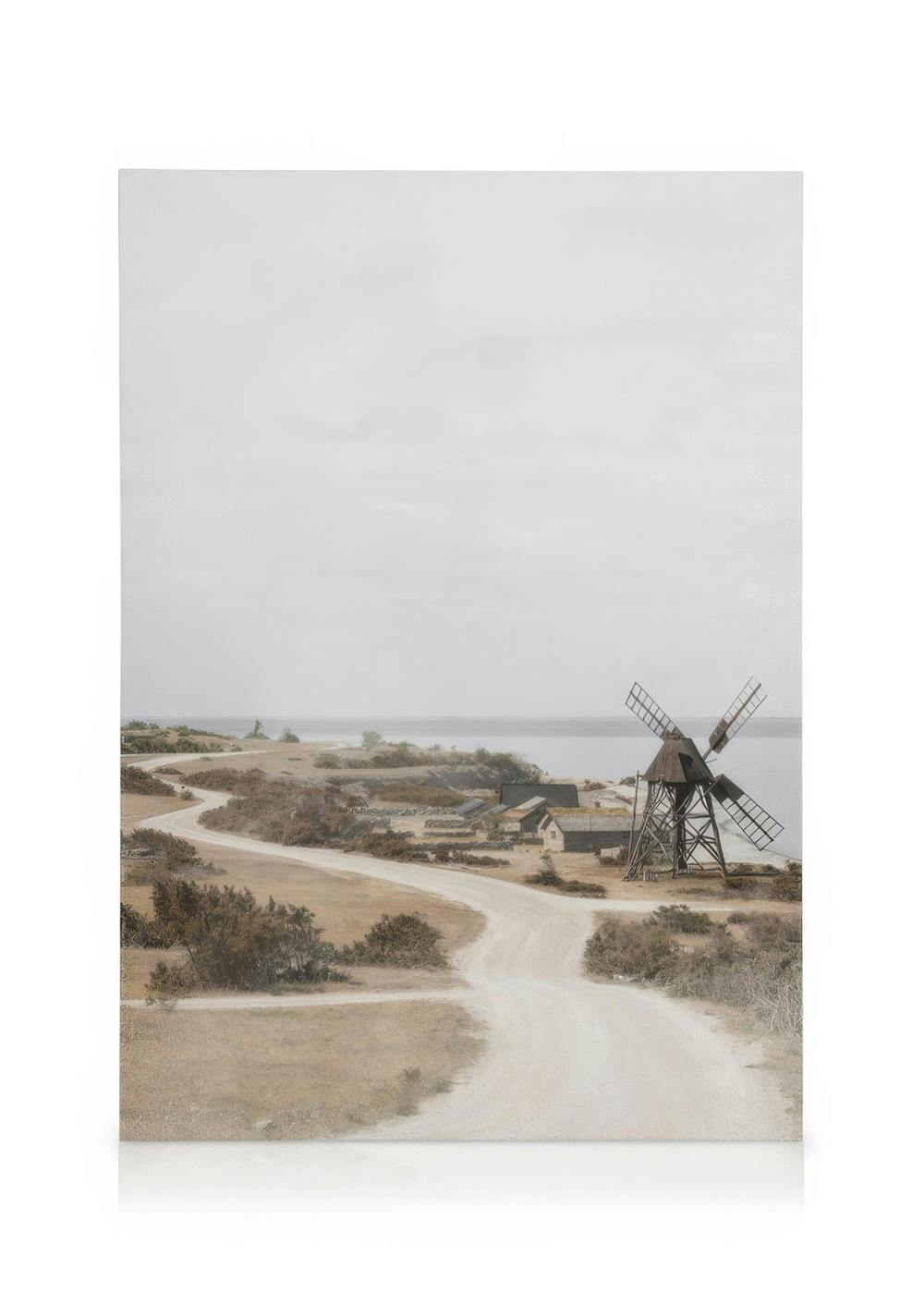 Obraz na płótnie: jałowy krajobraz z krętą drogą, starym wiatrakiem, krzewami i morzem pod pochmurnym niebem.