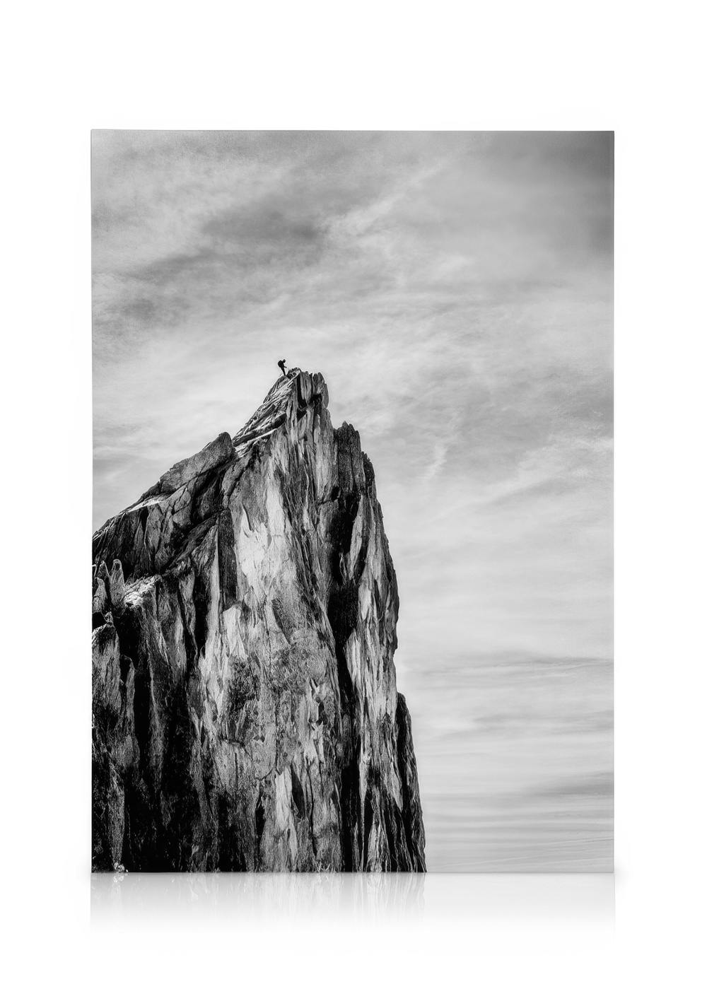 Black and white photography of a solitary figure standing atop a jagged mountain peak, reaching for the sky.
