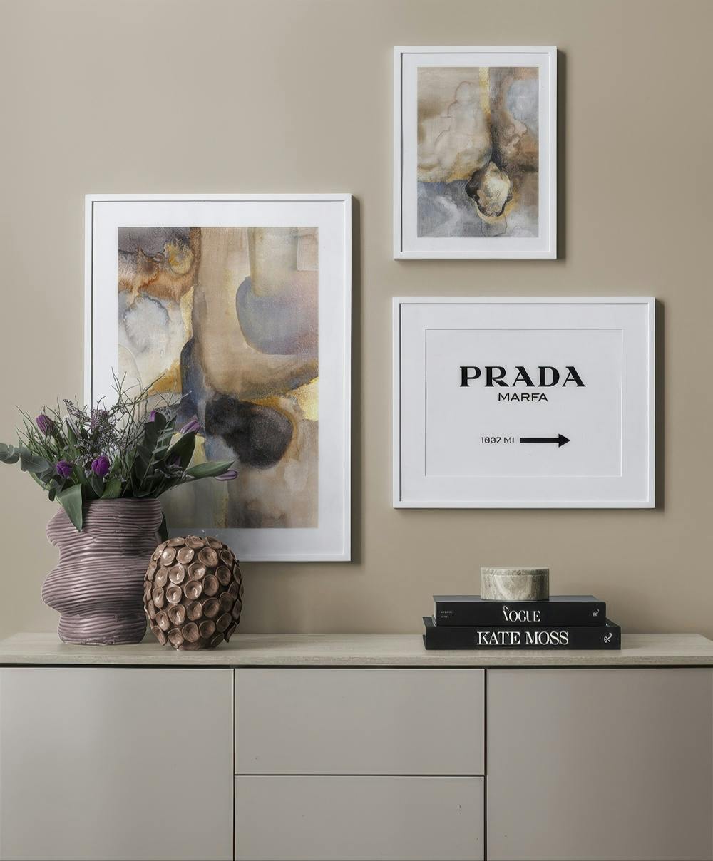 En tavelvägg med två abstrakta tavlor i beige och brunt samt en svartvit med texten PRADA MARFA.