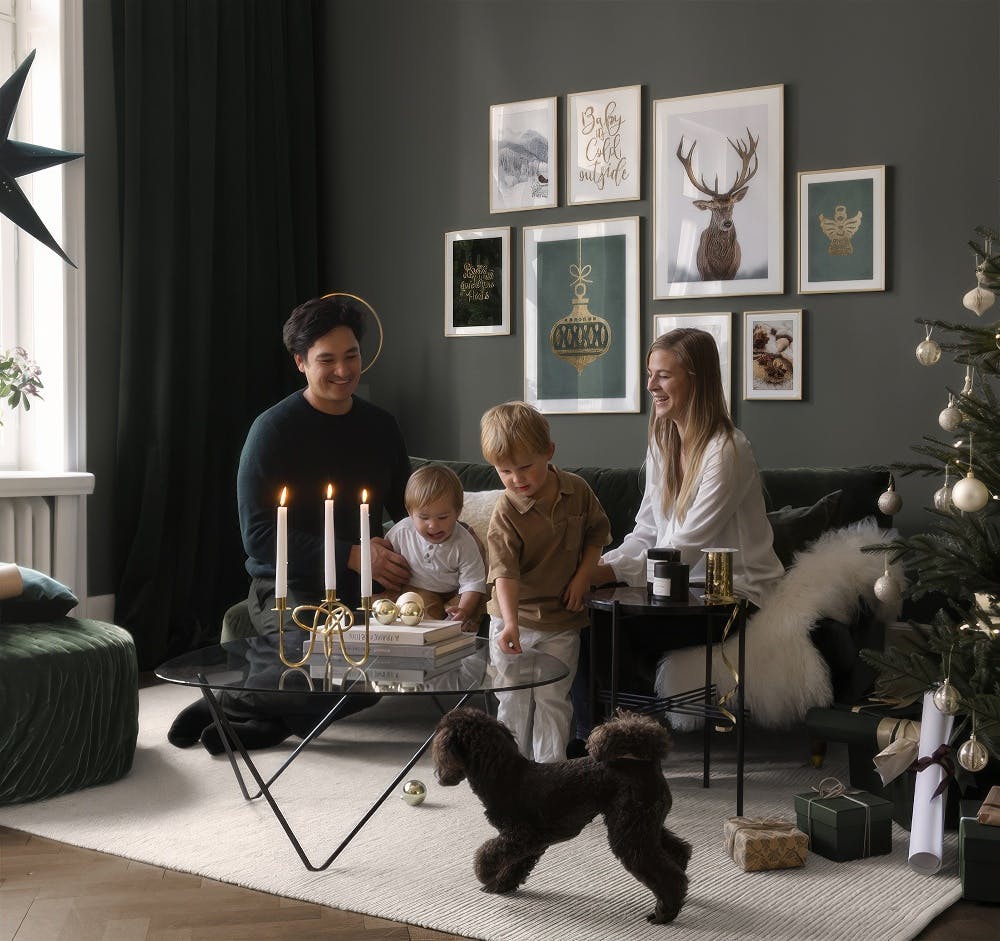 En tavelvägg med julmotiv i ett vardagsrum med en familj och en hund. Motiven inkluderar en kronhjort, en julkula, en ängel och