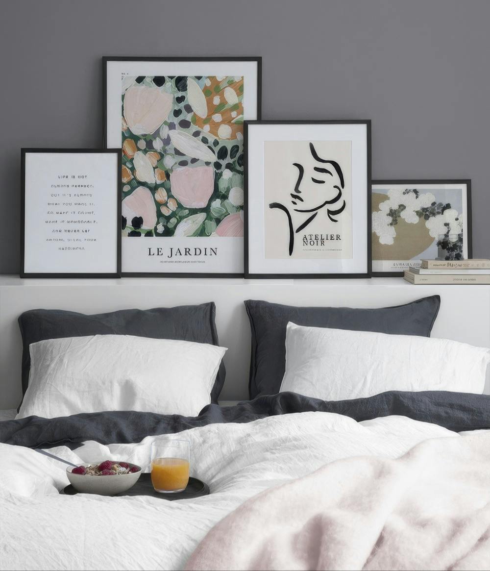 En tavelvägg med fyra posters ovanför en säng. Posters med text, abstrakt blommotiv, ansikte och landskap.