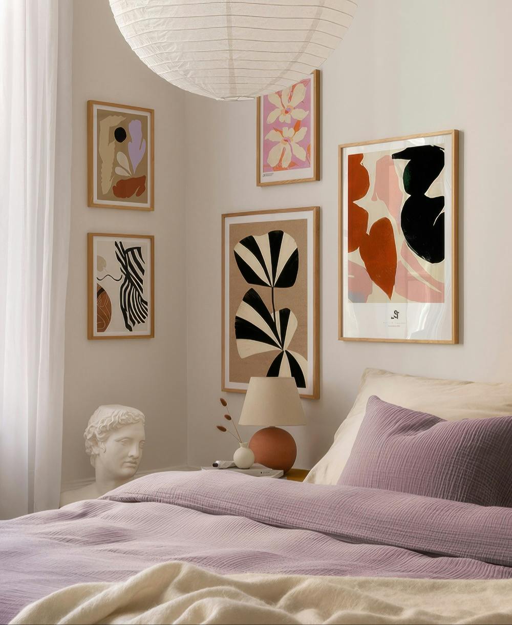 En tavelvägg med abstrakta botaniska motiv i varma färger som brunt, beige, svart, rosa och lila i ett sovrum.