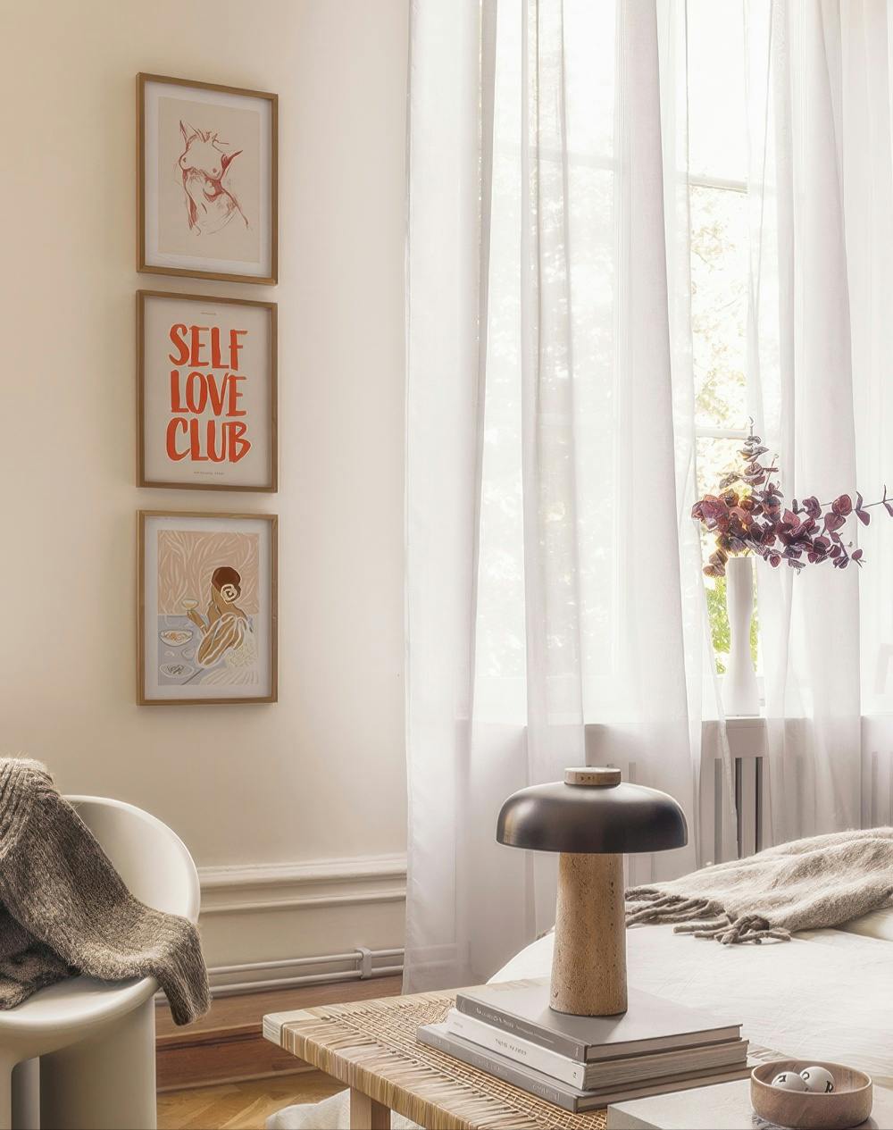 Een galeriewand met drie prints: een contourtekening van een vrouwenlichaam, rode tekst SELF LOVE CLUB en een vrouw aan tafel.