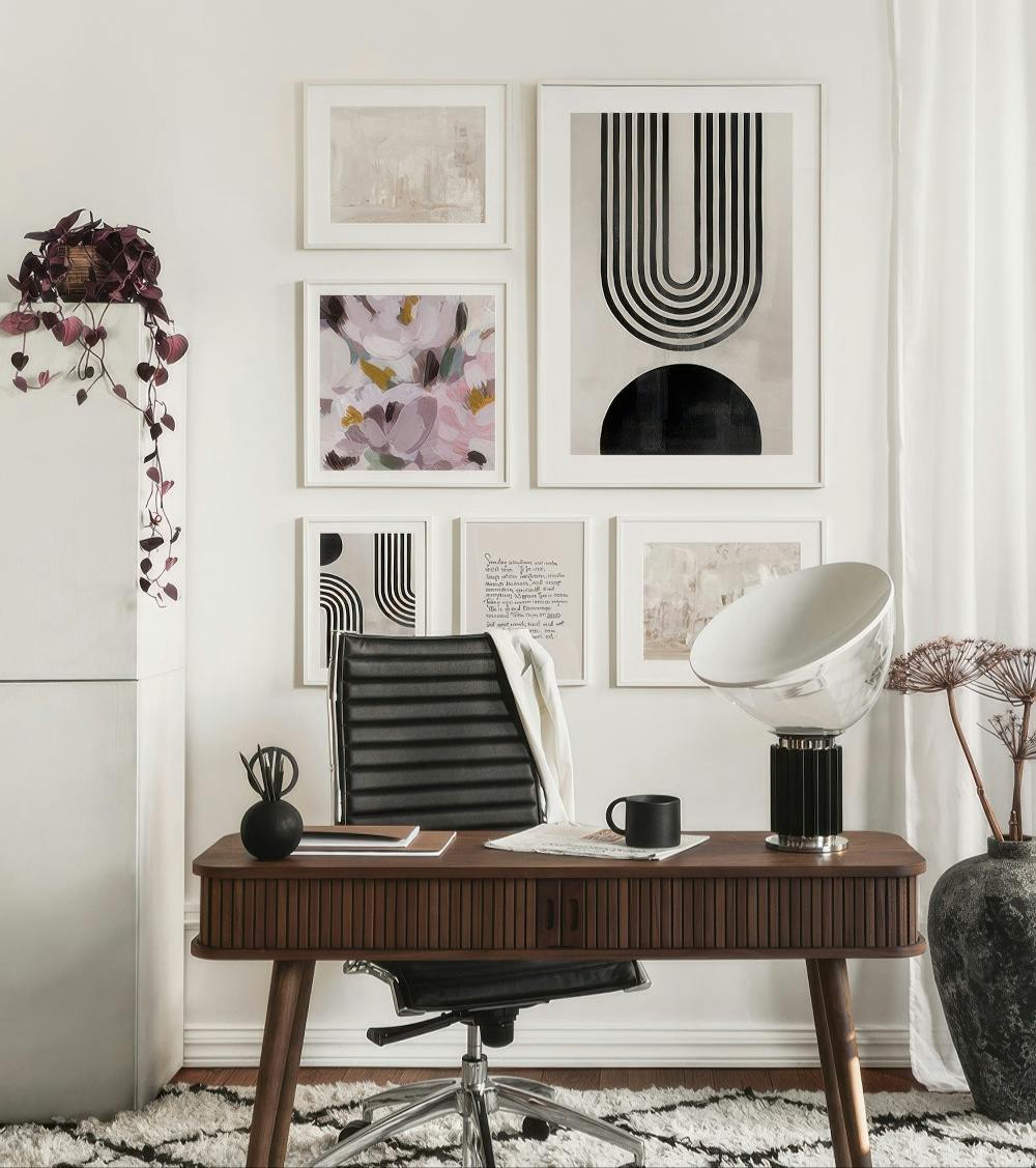 En billedvæg med kunst i jordfarver og pasteller over et skrivebord, inkl. en abstrakt bue og blomster.