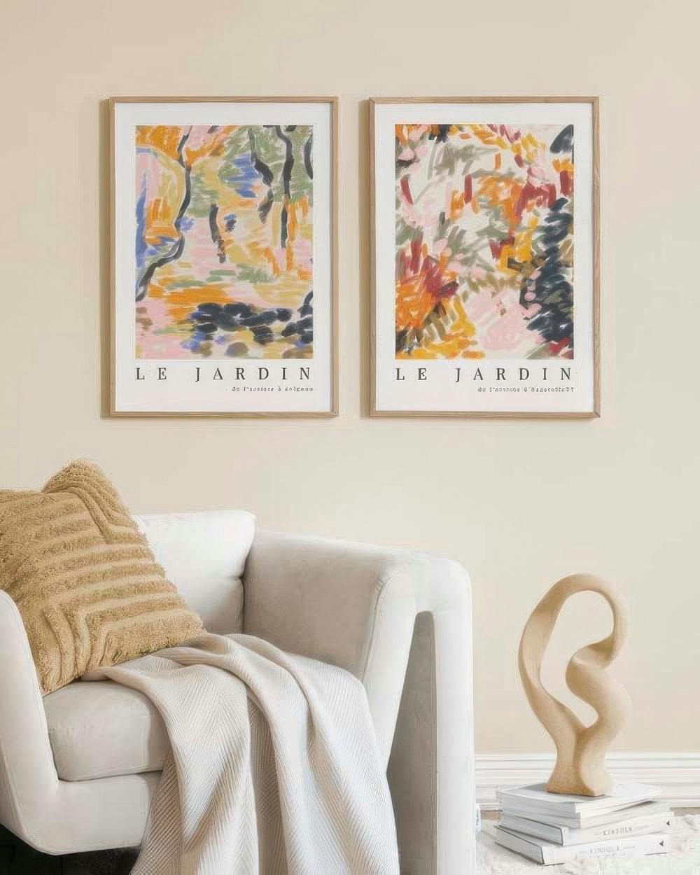 En tavelvägg med två abstrakta posters i varma färger som hänger över en ljus fåtölj och en beige skulptur.