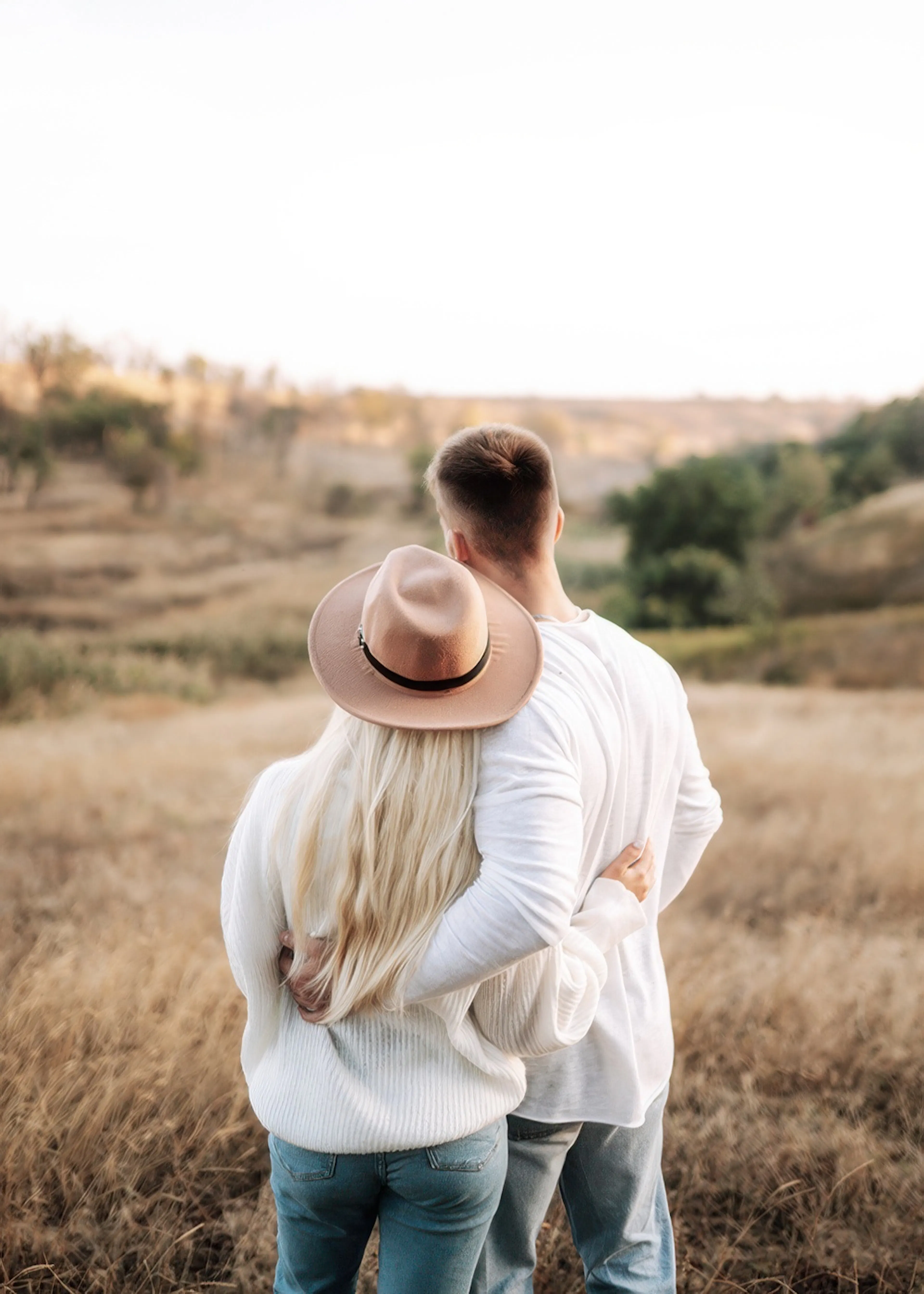 Paar umarmt sich von hinten auf einem Feld, Frau trägt Hut, goldene Stunde, Natur, Liebe