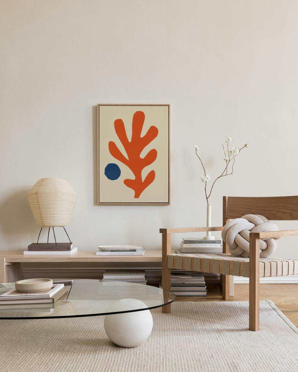 Abstract canvas print with an orange leaf shape and blue dot on a light background, hanging in a modern living room.