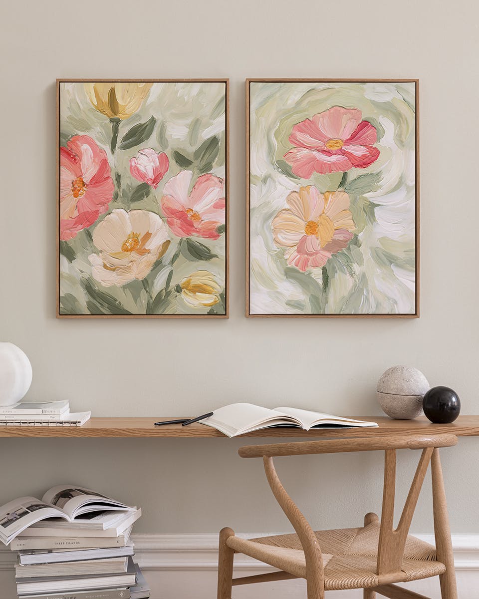 Two floral prints with pink and yellow flowers on a green and white background, displayed above a wooden desk with books and a c