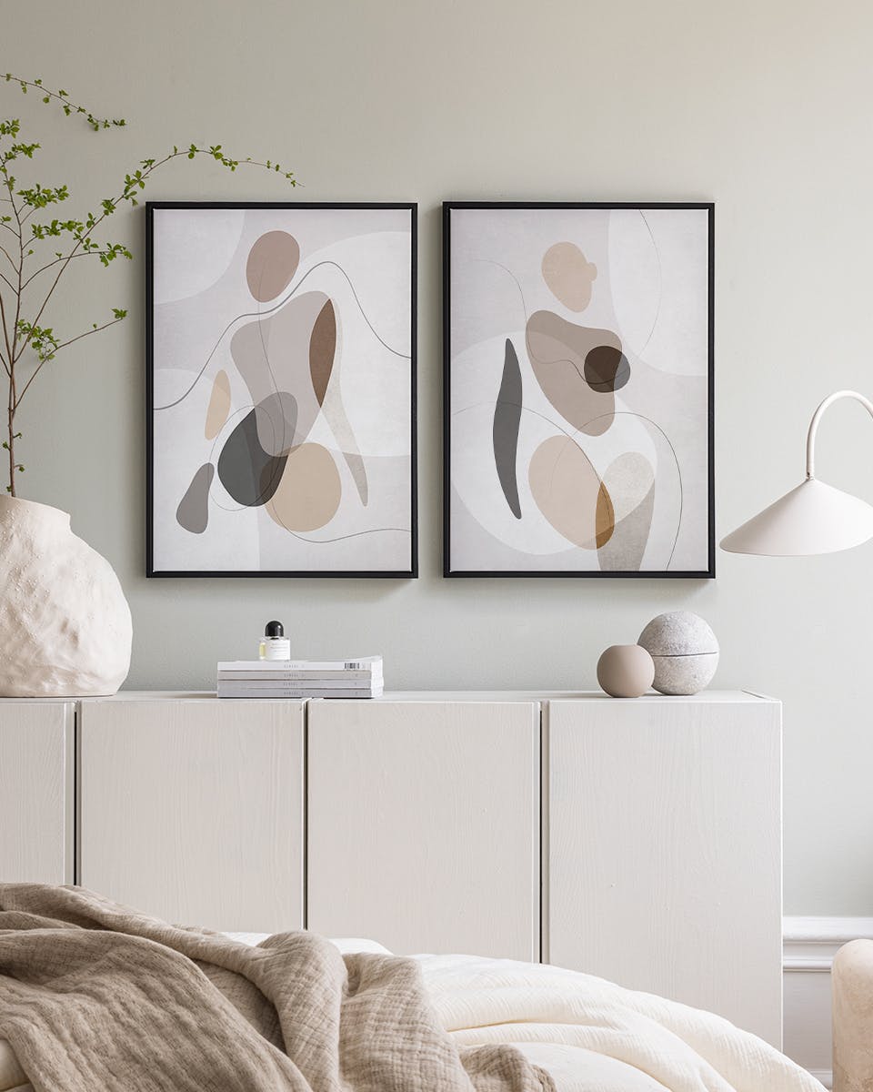 Two abstract poster prints with organic shapes in beige, brown, and grey, hanging above a white console table in a bedroom.