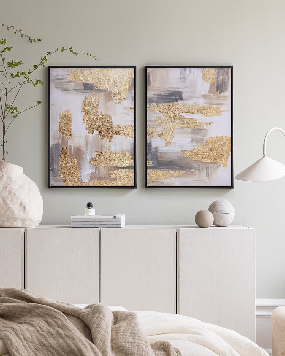 Two abstract poster packs with gold, grey, and white brushstrokes, displayed above a white cabinet in a bright room.