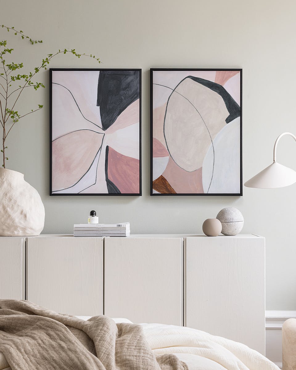A poster pack featuring two abstract prints with organic shapes in pink, black, and beige, displayed above a white cabinet in a
