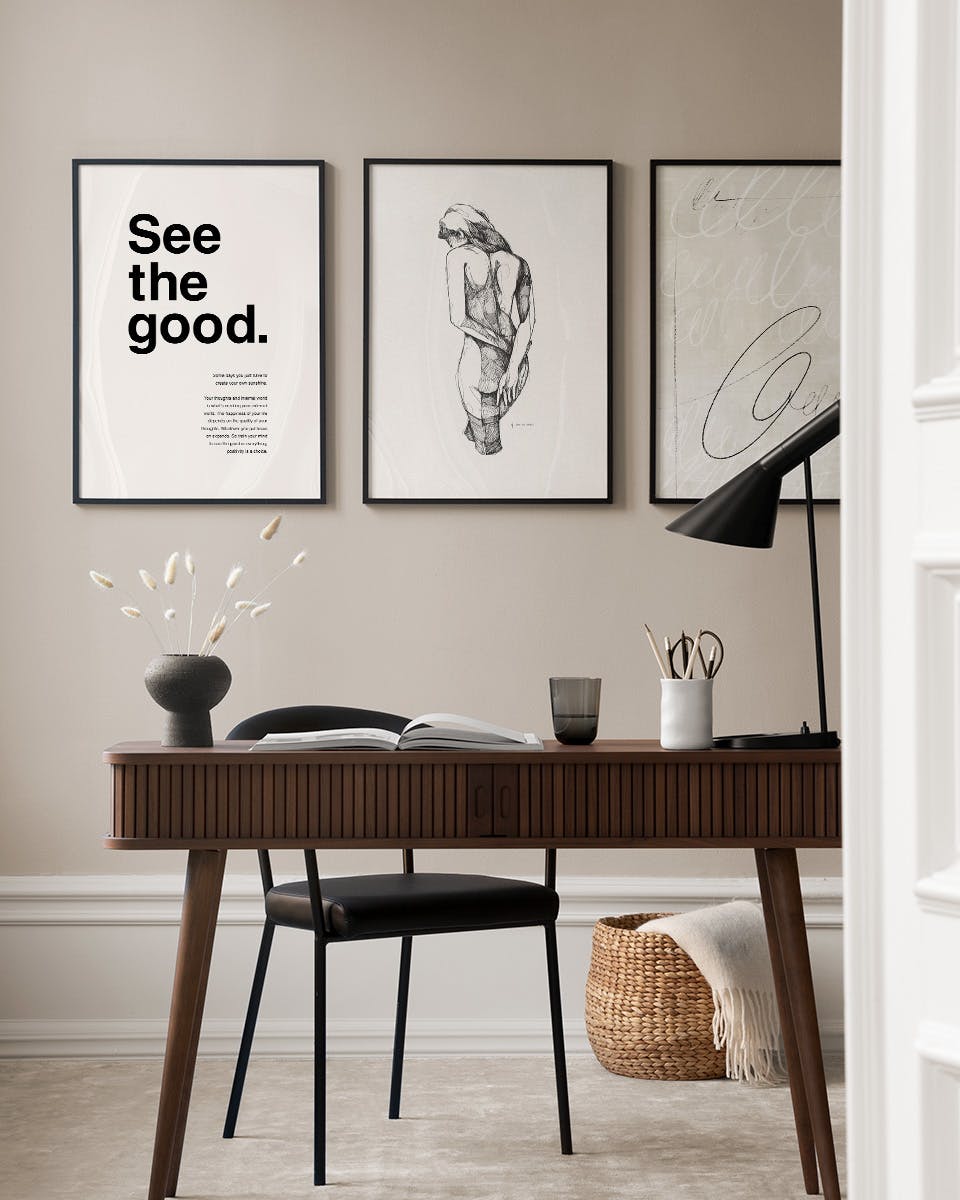 A gallery wall of three prints above a wooden desk. One features See the good. text, another a nude figure drawing, and the thir