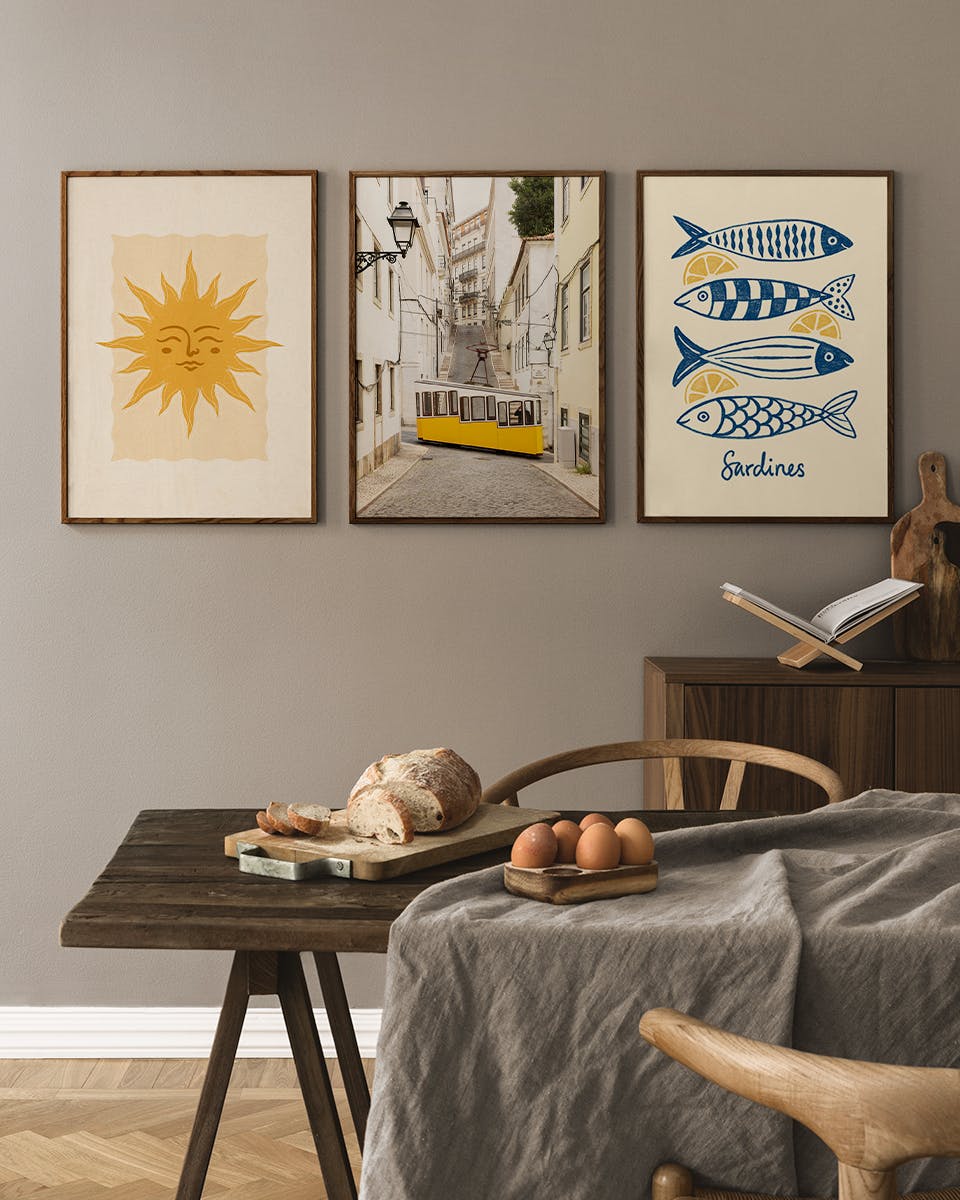 Plakatpakke med tre plakater: en sol, en gul sporvogn i Lissabon og sardiner, hængende over et træbord med brød.