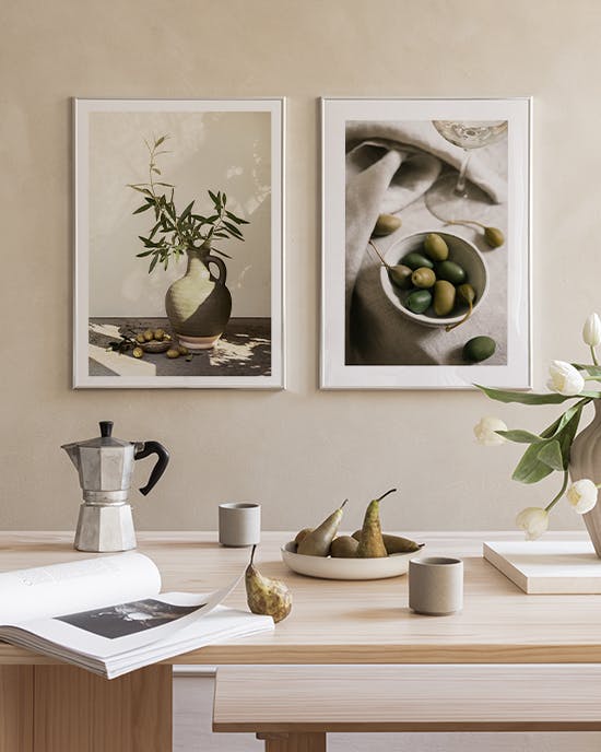 Två naturbilder i gråskala, en med olivkvistar i vas och en med oliver, hänger ovanför ett träbord med kaffekanna och päron.