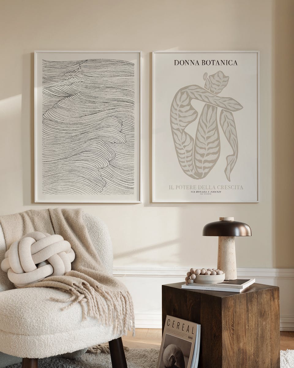 Two poster prints on a cream wall, one with abstract black wavy lines, the other with a beige figure, beside a plush chair and w