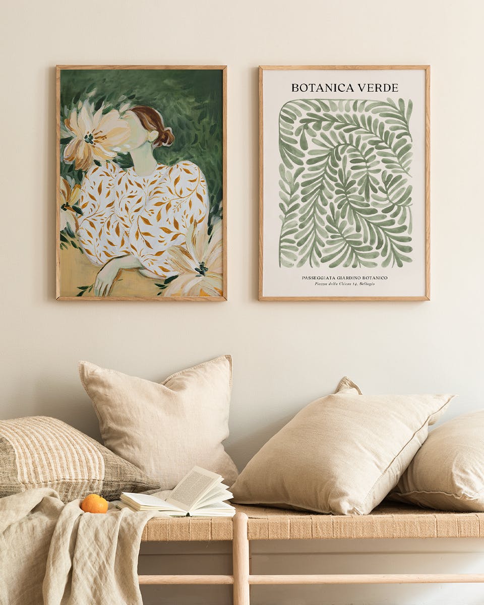 Two botanical art prints hanging above a light wood bench with neutral pillows and an open book.