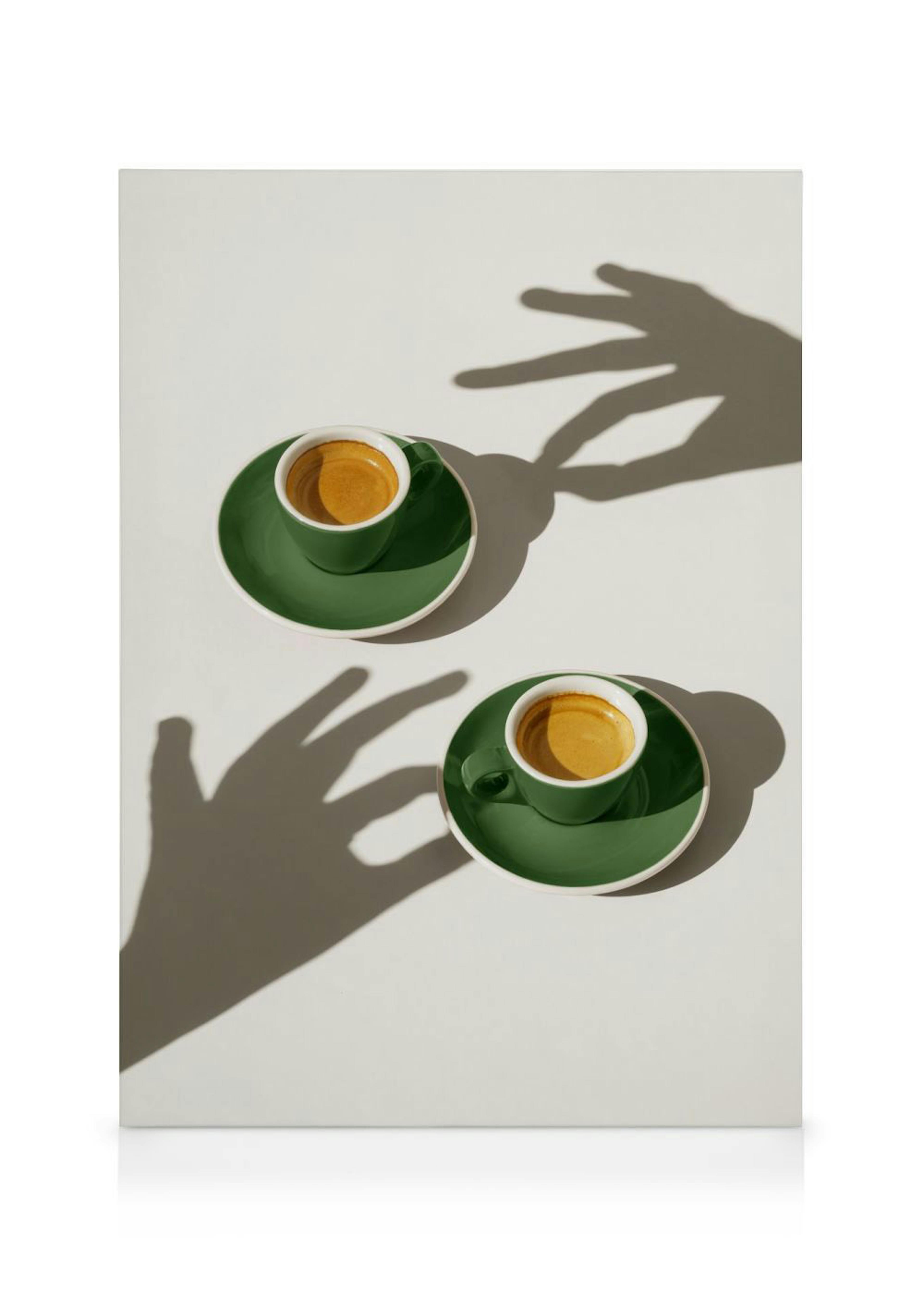 A canvas print featuring two green espresso cups with saucers, casting shadows of hands on a white surface.