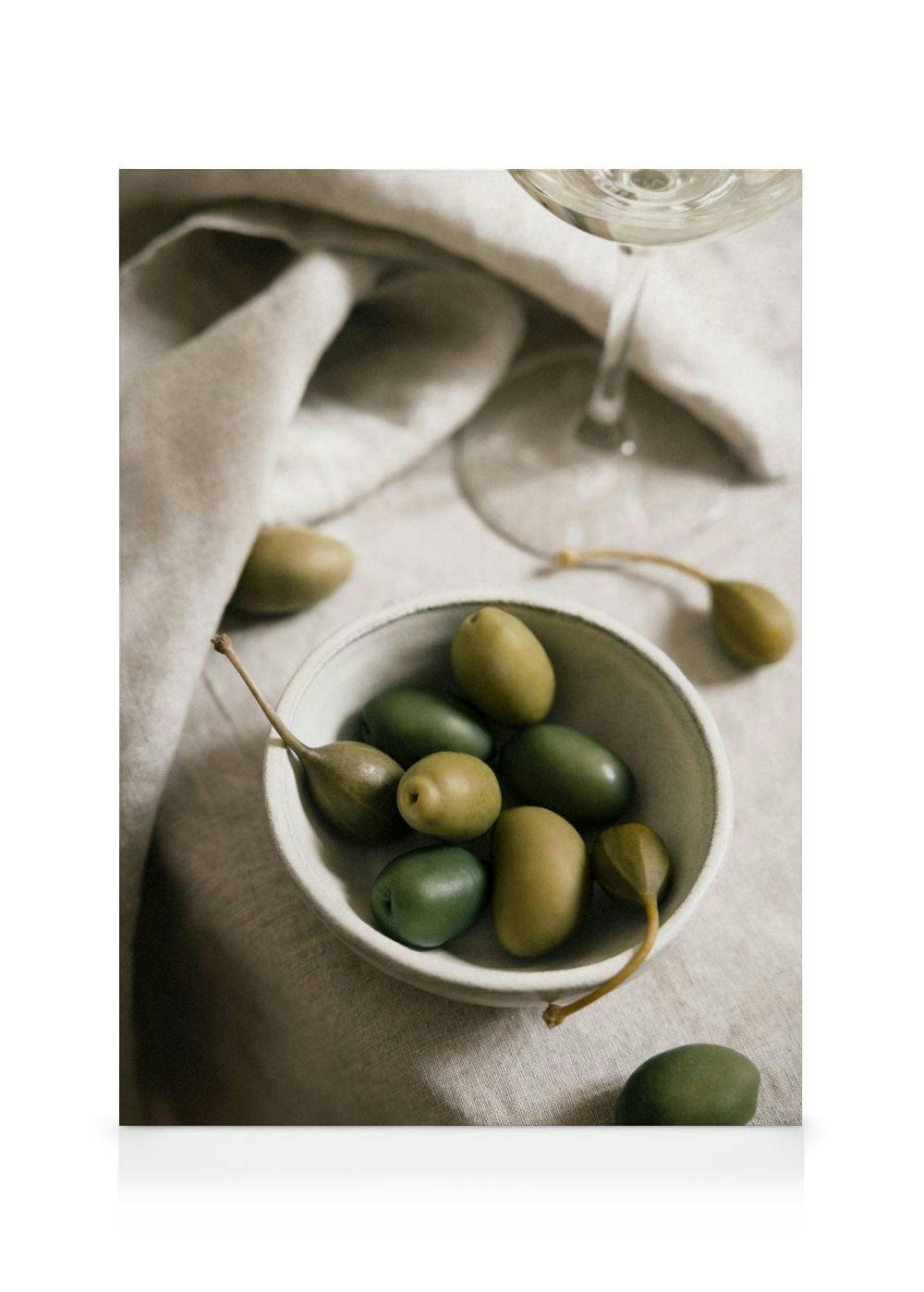 A canvas print featuring a bowl of green olives and caper berries on a light linen tablecloth, with a wine glass in the backgrou