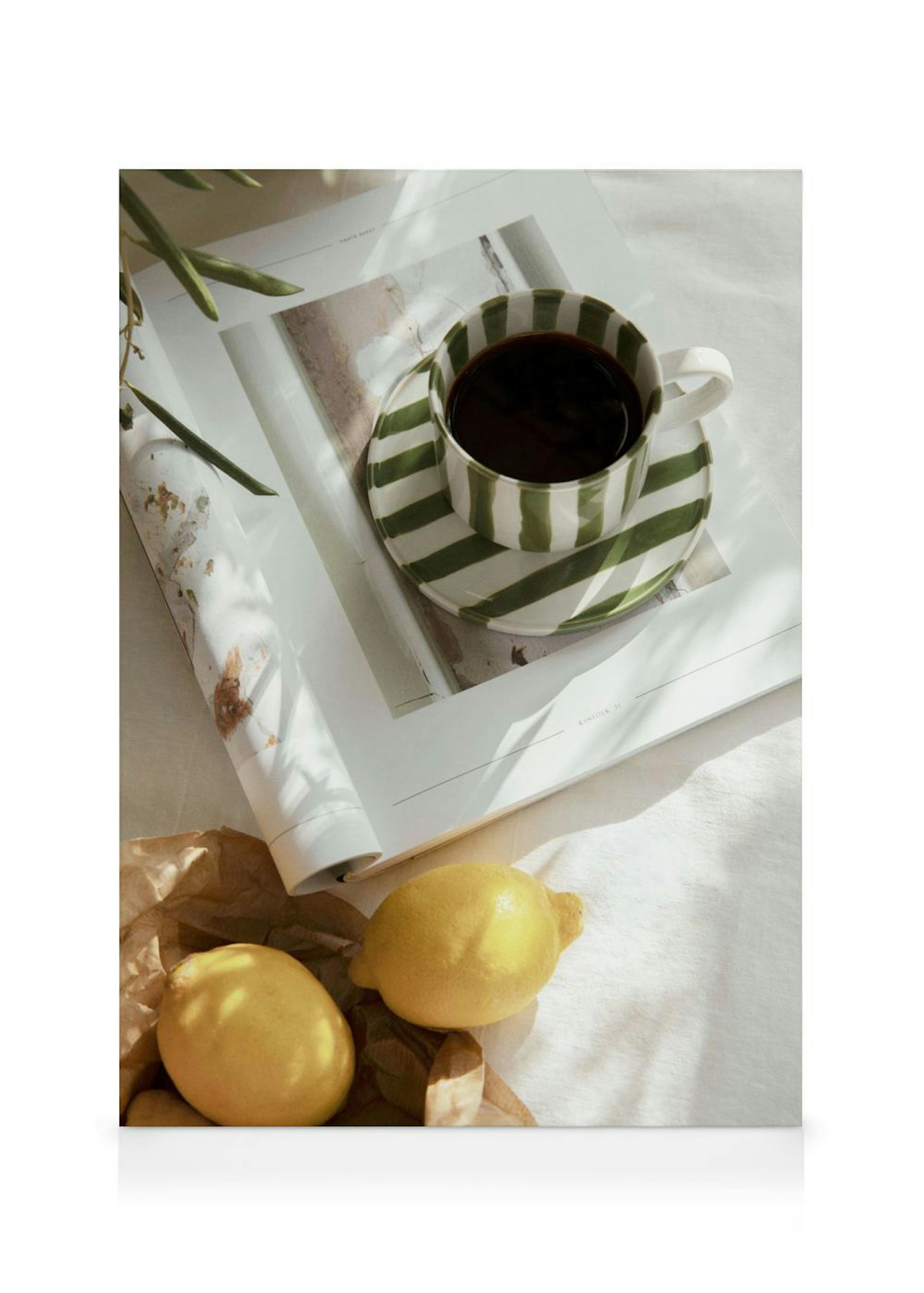 En canvastavla med en grönrandig kaffekopp på ett magasin, tillsammans med två citroner på en vit duk.