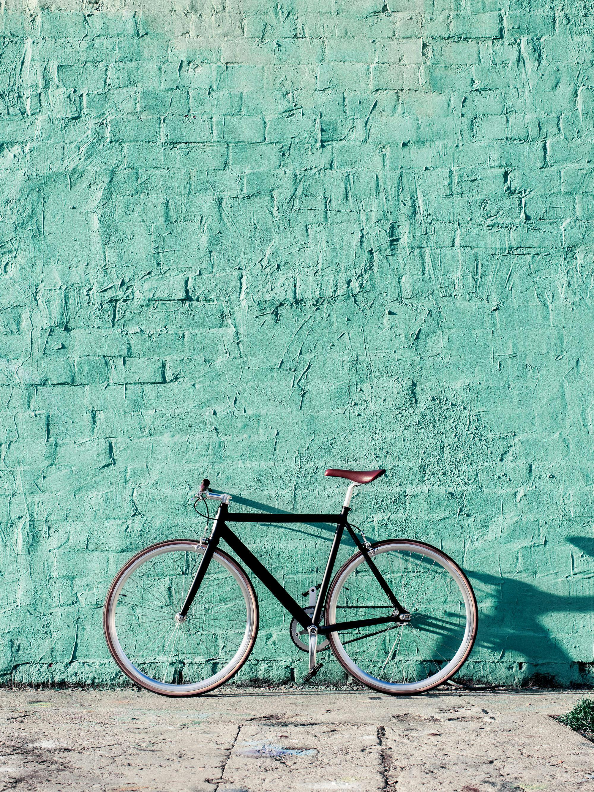 Svart cykel med brunt säte mot mintgrön tegelvägg och grå betongtrottoar, fotokonstposter.