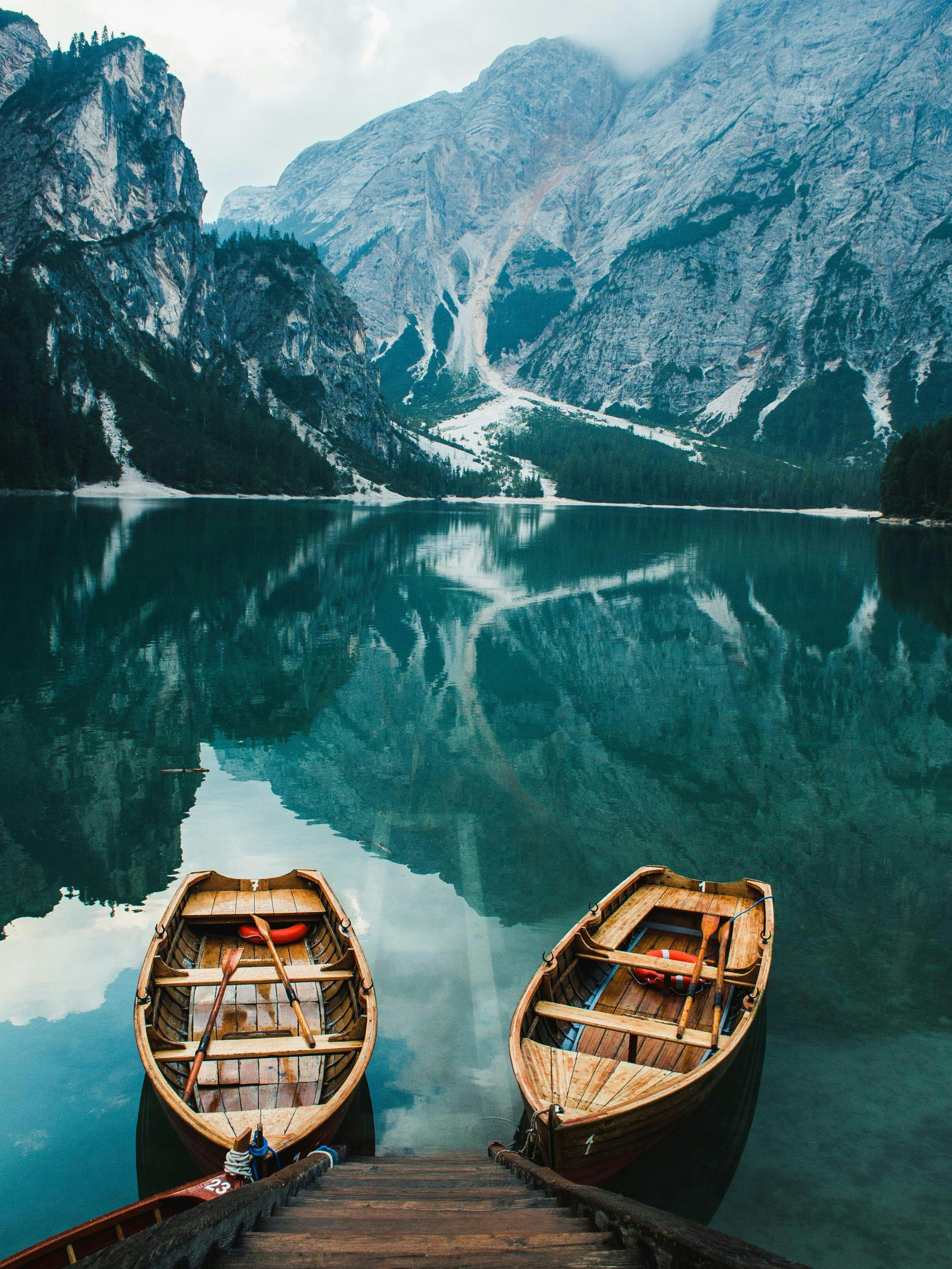 Fotografi av två träbåtar på en lugn sjö, omgiven av dimmiga berg och skog. Naturlandskap.