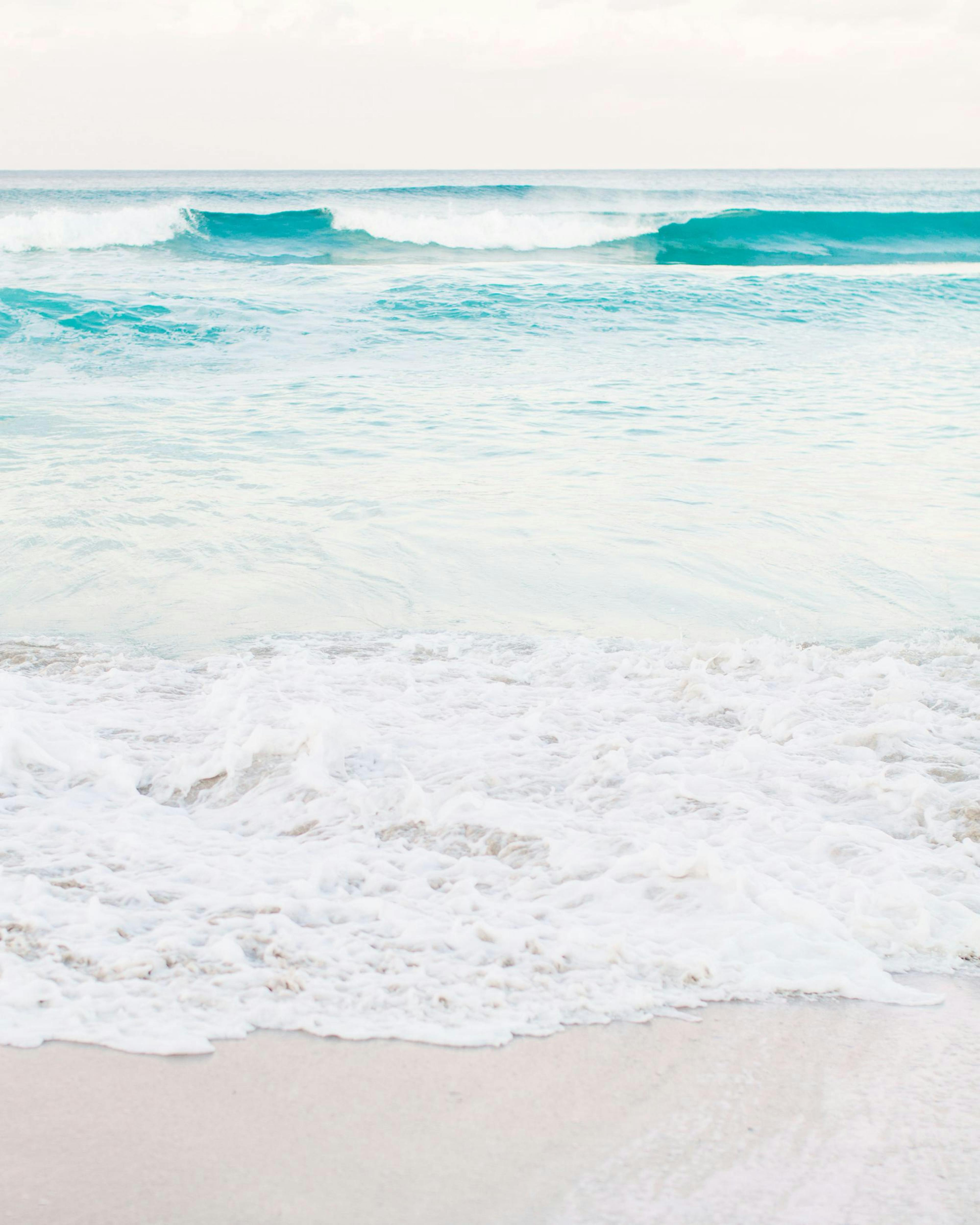 Pastel photo of teal ocean waves crashing on a sandy beach shore with white seafoam