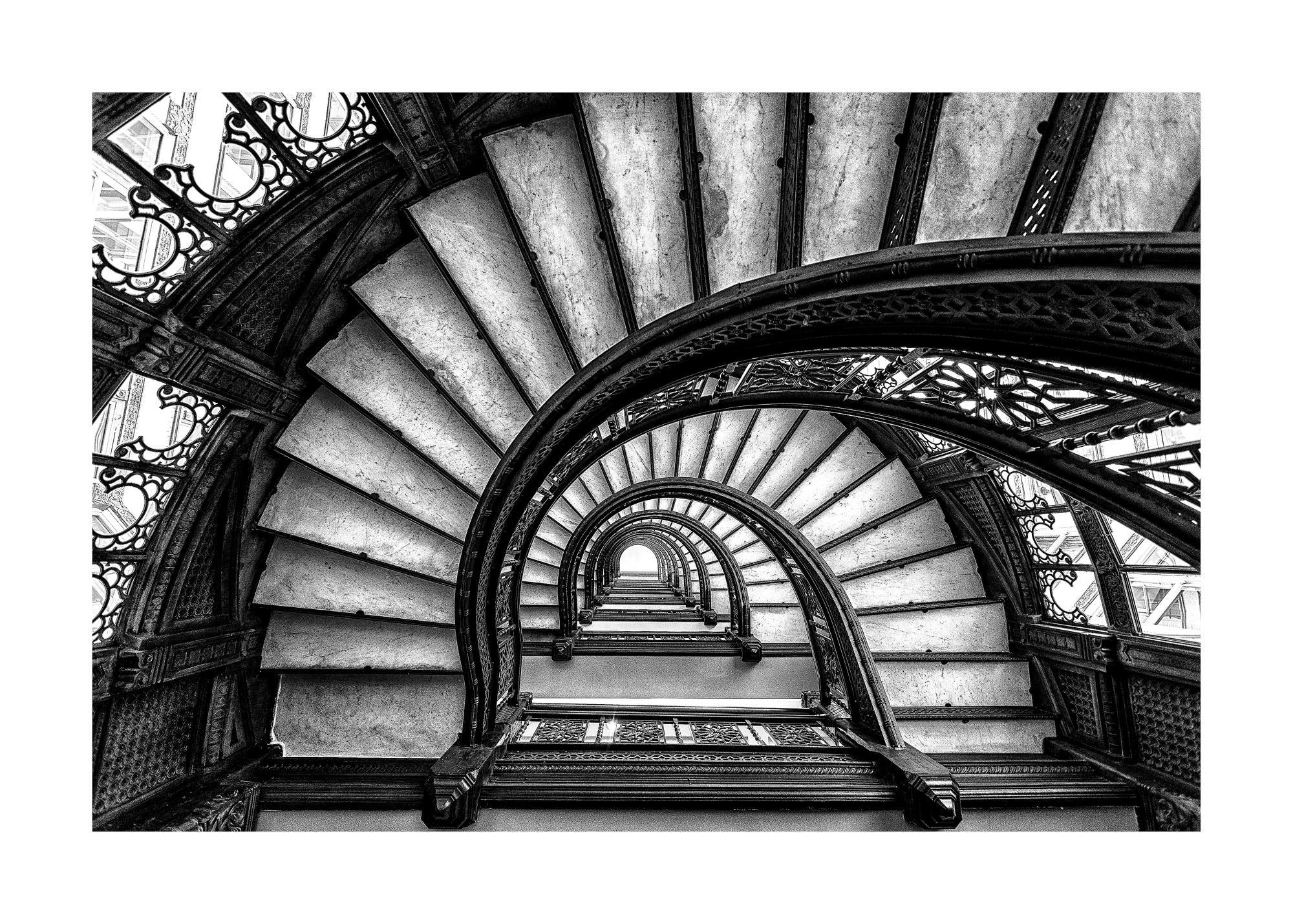 Black and white poster print of a circular spiral staircase viewed from above, featuring ornate wrought iron railings.