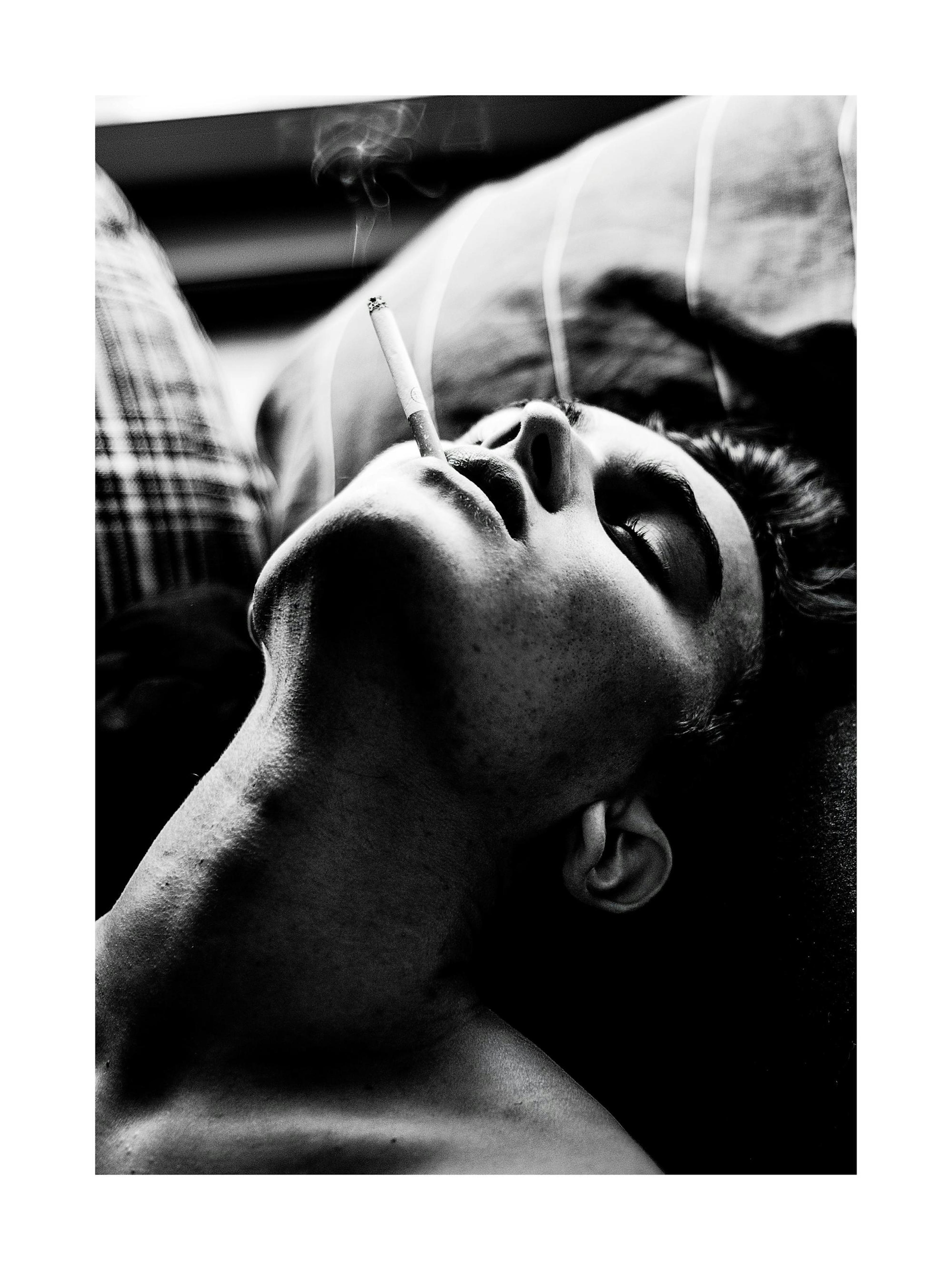 Black and white print of a person smoking in bed, looking up with smoke rising from their cigarette.