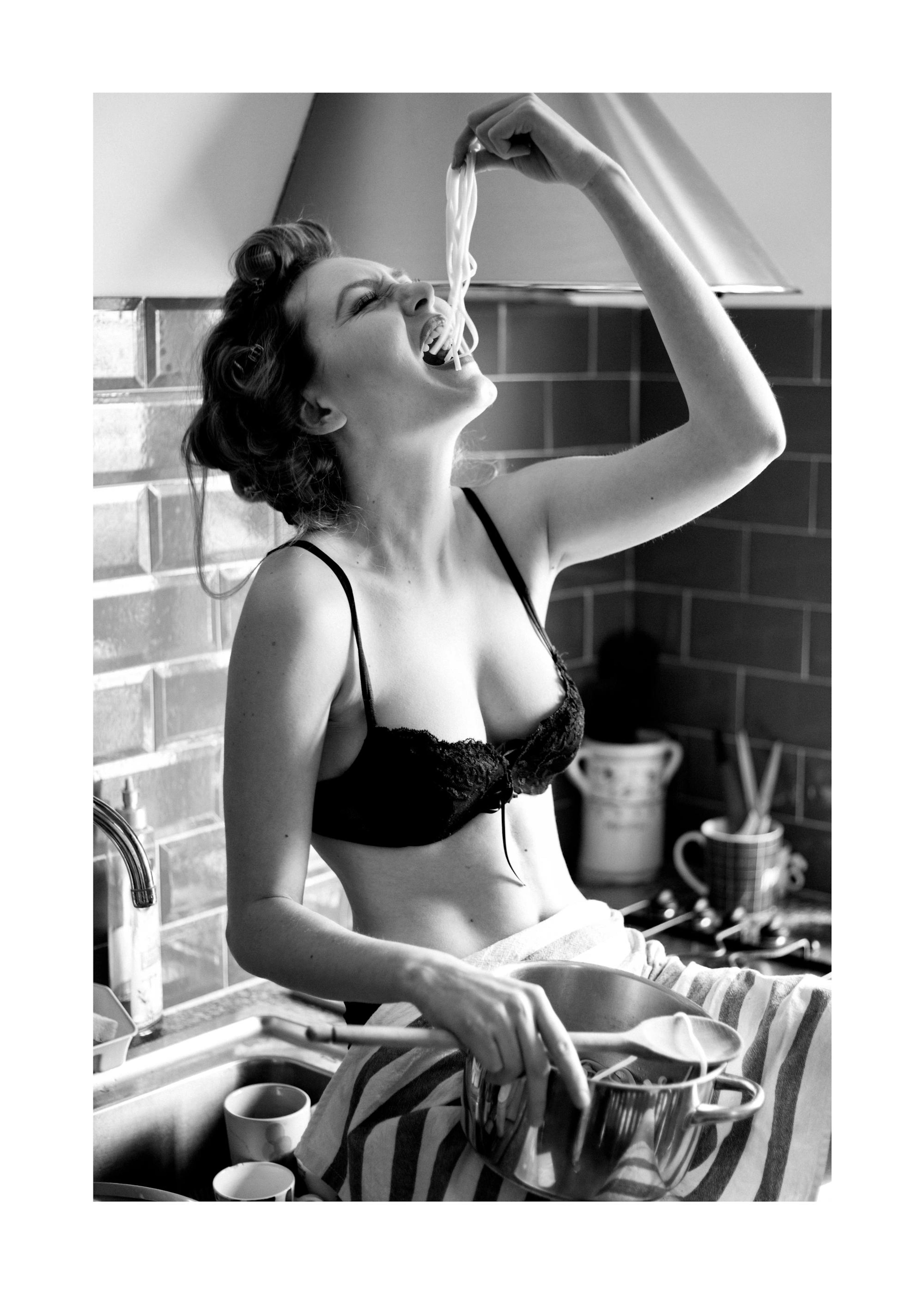 Black and white photograph of a woman in a bra eating pasta in a kitchen, featured on a print poster.