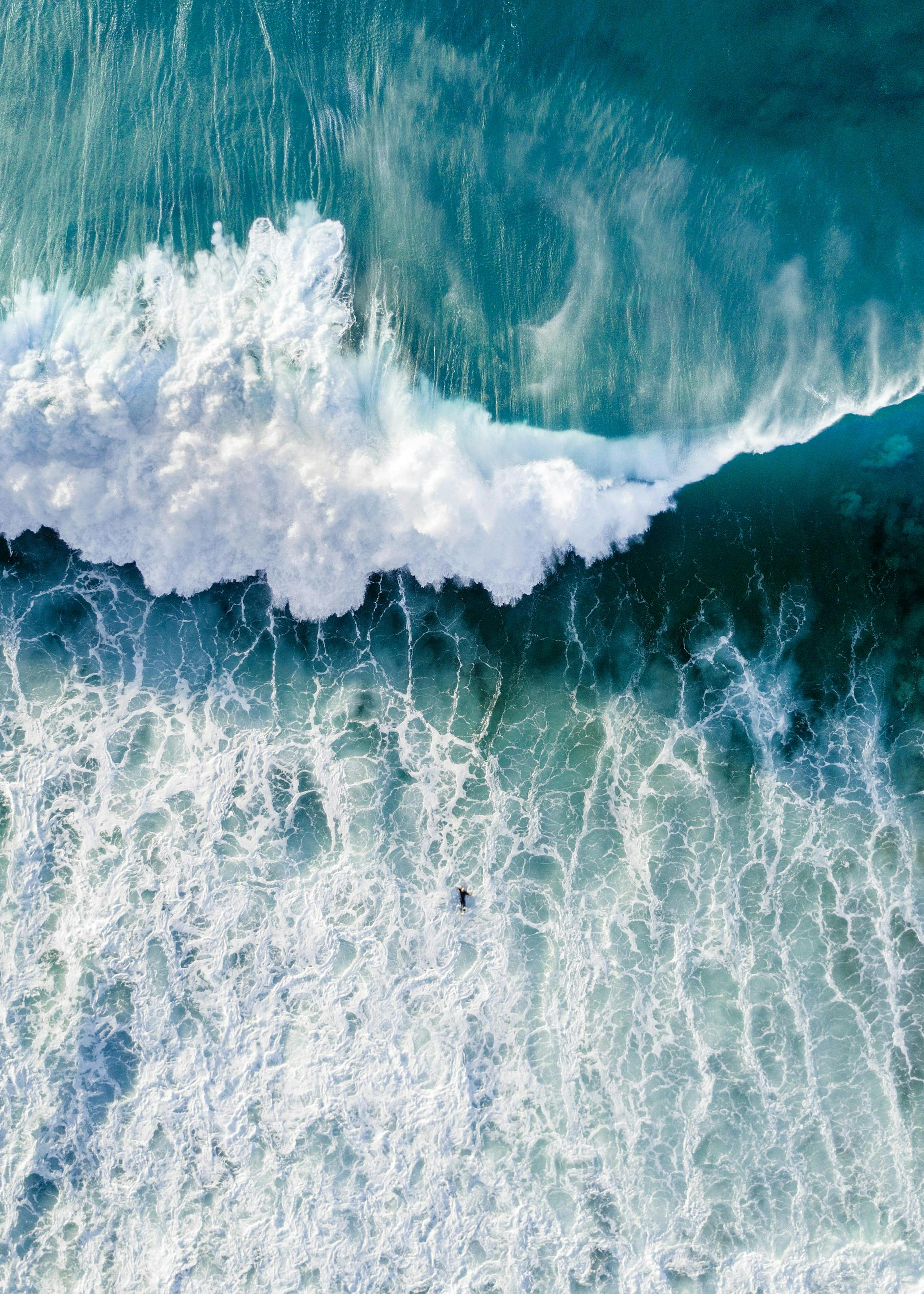 Aerial photograph of a powerful ocean wave crashing with white foam, a lone surfer visible in the turquoise water.