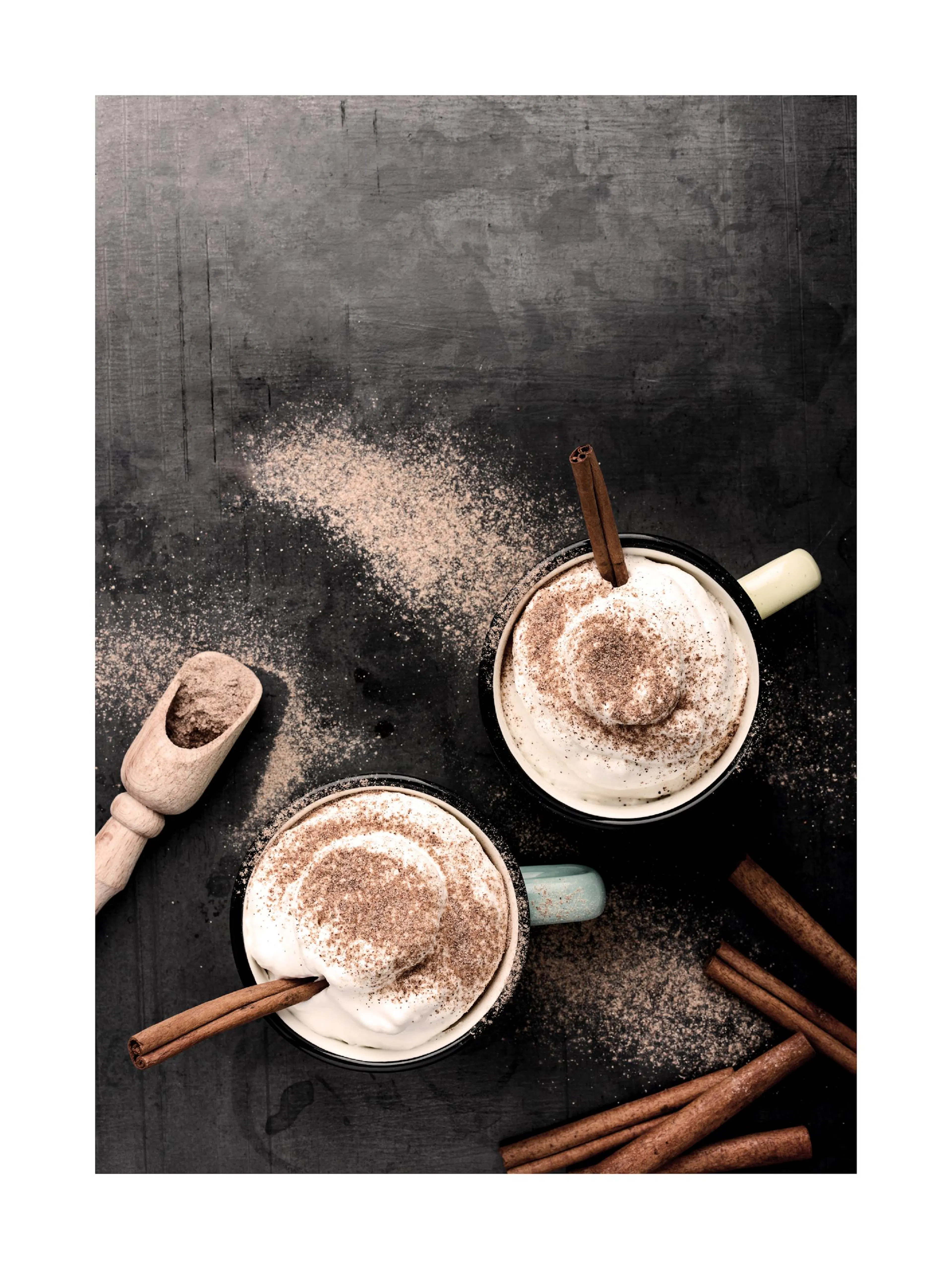 Overhead view of two caffè latte mugs with whipped cream, cinnamon sticks, and sprinkled cocoa on a dark background print.