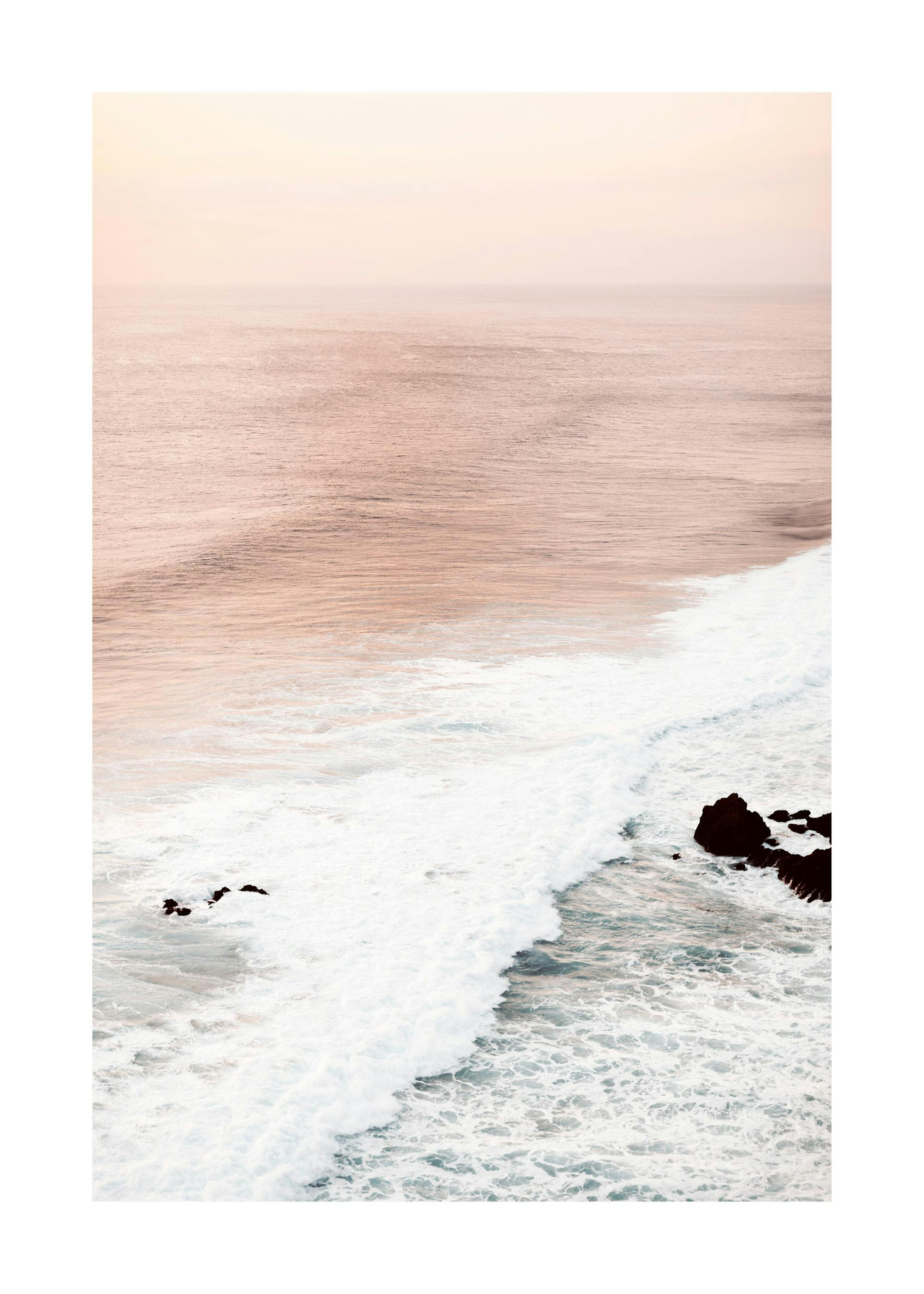 Soft Waves Print: Aerial photograph of a pink ocean with white waves crashing on rocks under a light pink sky.