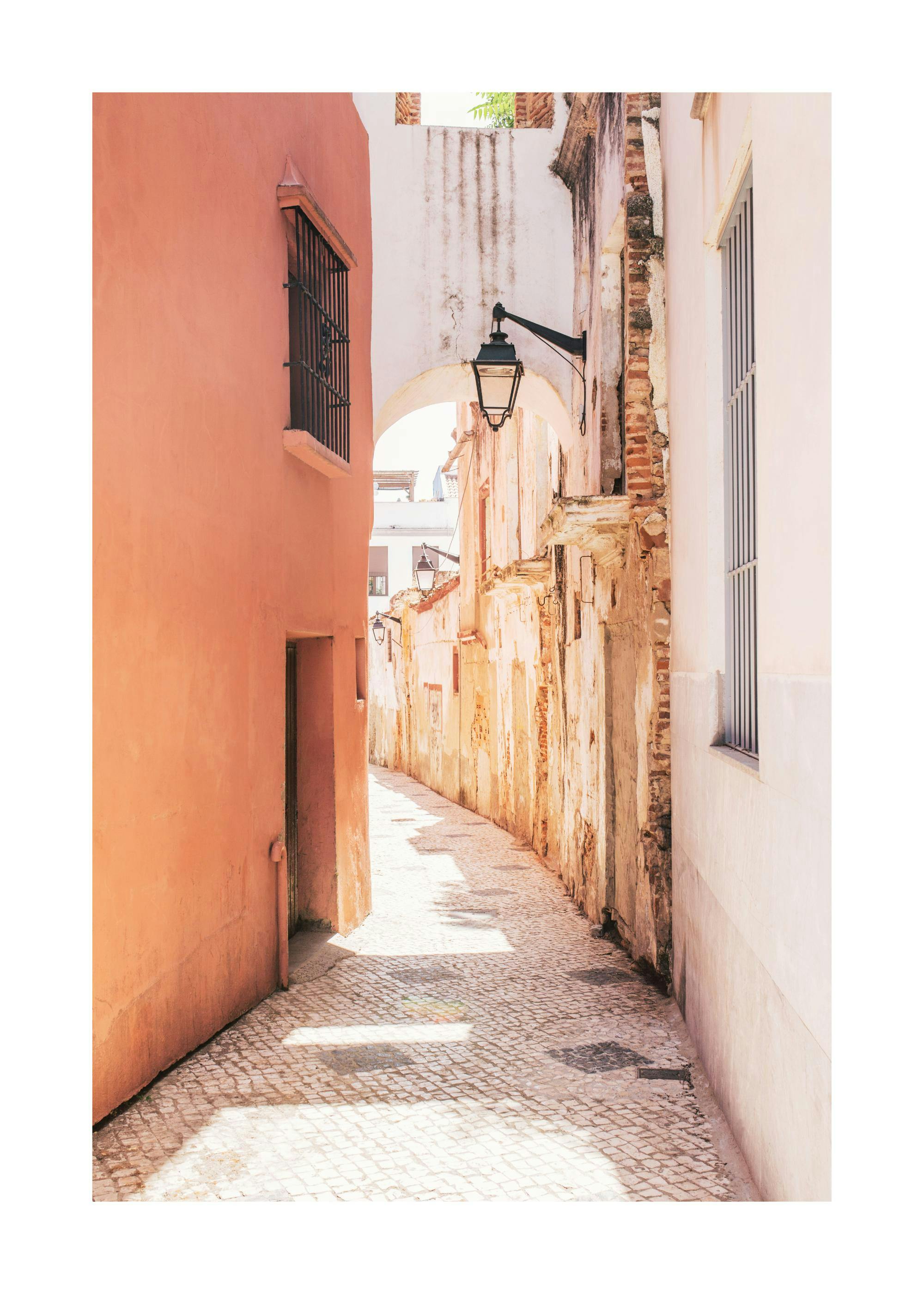 Spanish alley print with narrow cobblestone path, light pink and white buildings, and arched entryway with lantern