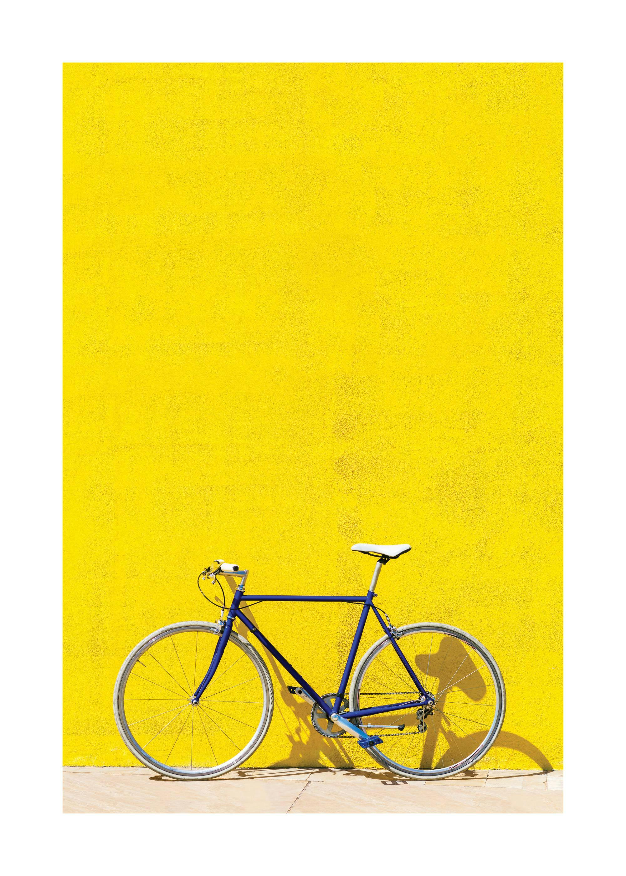 Blue bike with white seat and handlebars against a vivid yellow wall, casting a shadow on the ground.