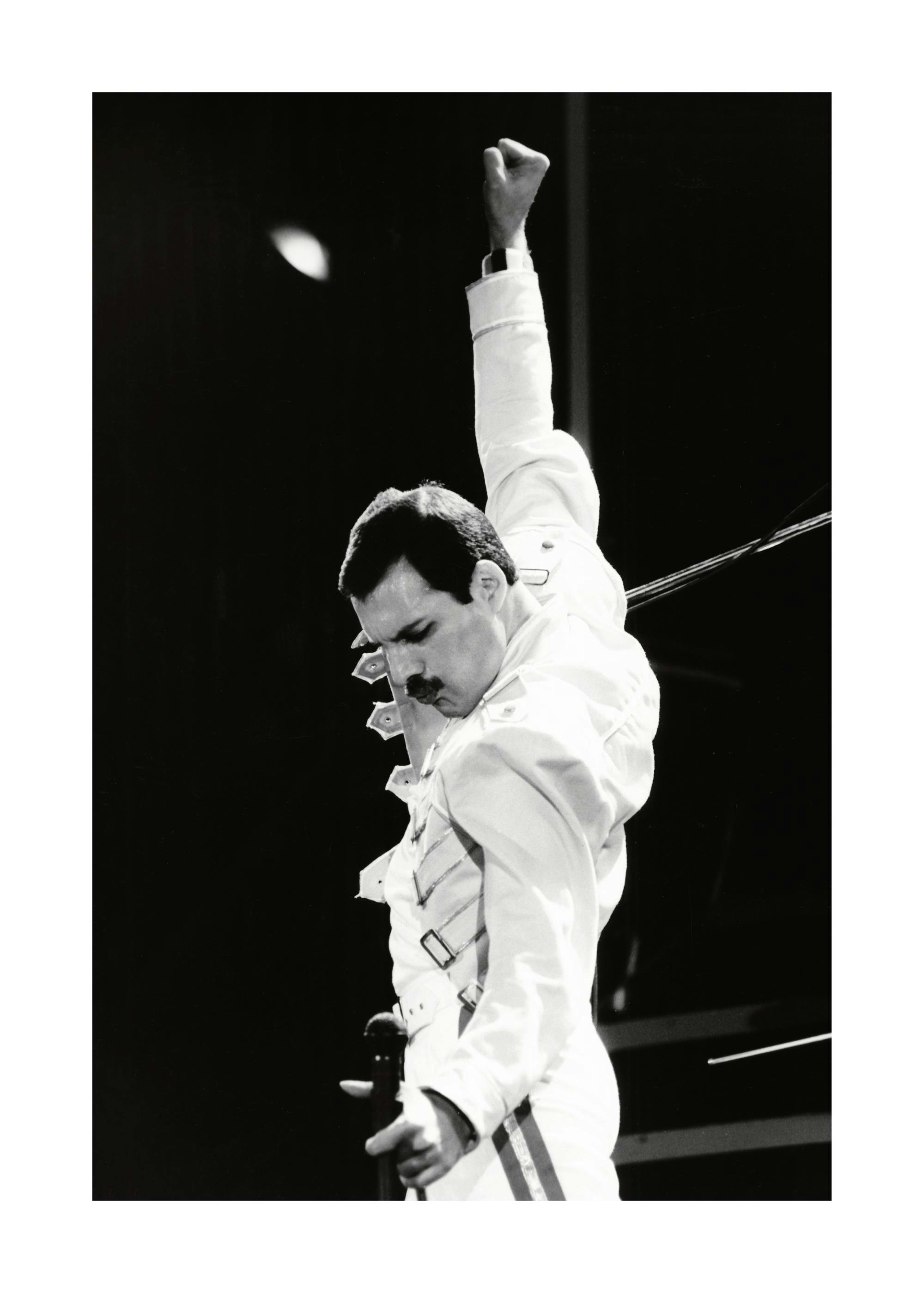 Fotografia in bianco e nero di Freddie Mercury dei Queen, vestito di bianco con un braccio alzato.