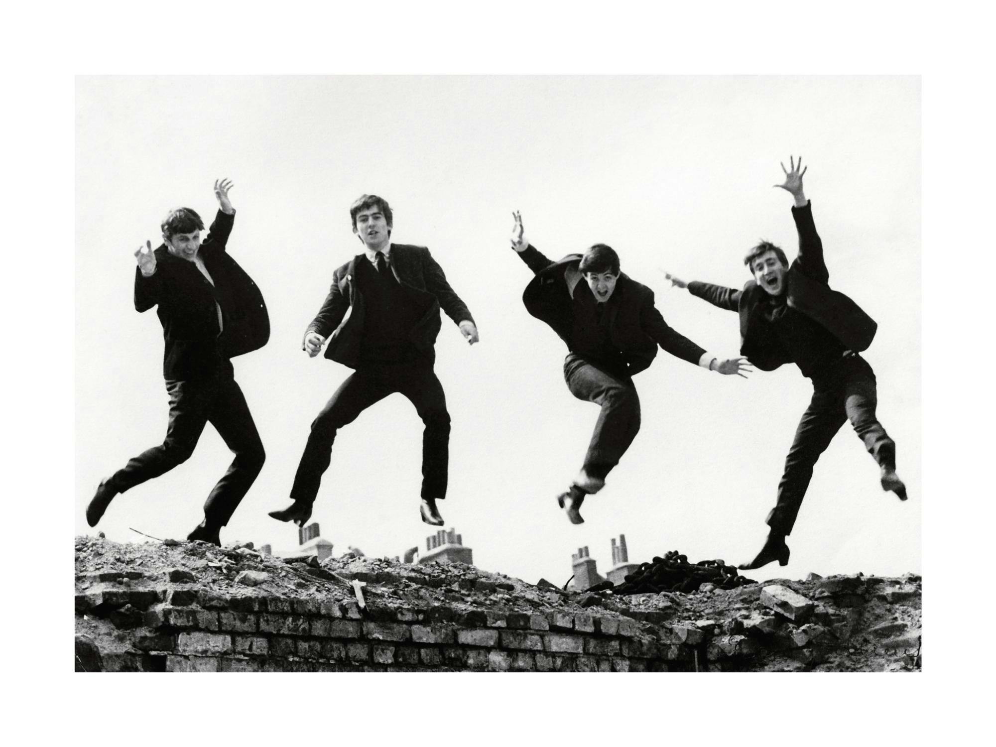Svartvitt fotografi av The Beatles som hoppar i luften över en raserad mur. Ikonisk bild av popgruppens medlemmar.
