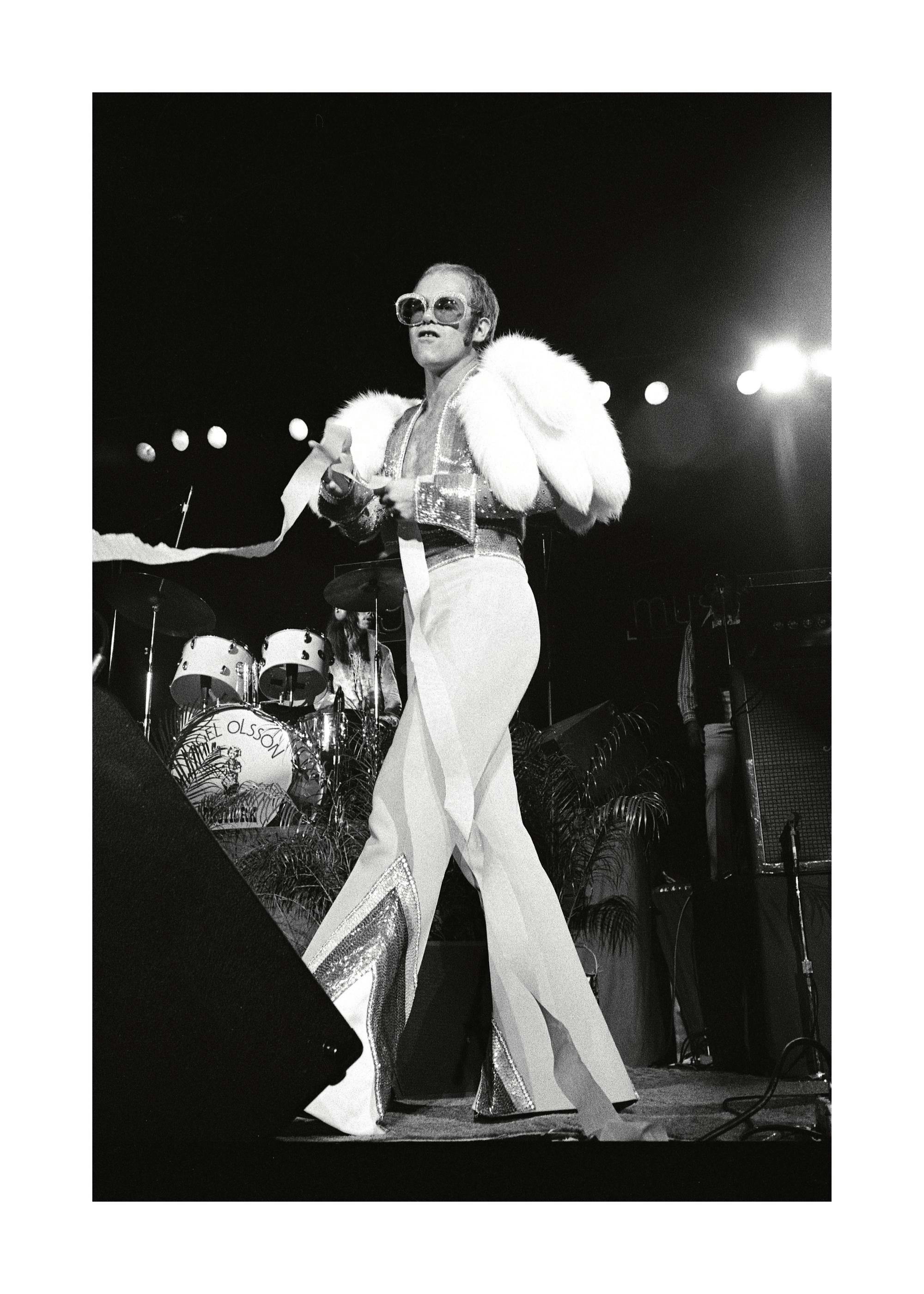 Fotografia a preto e branco de Elton John com óculos e casaco de penas em palco. Pôster musical icónico, estilo retro vintage.