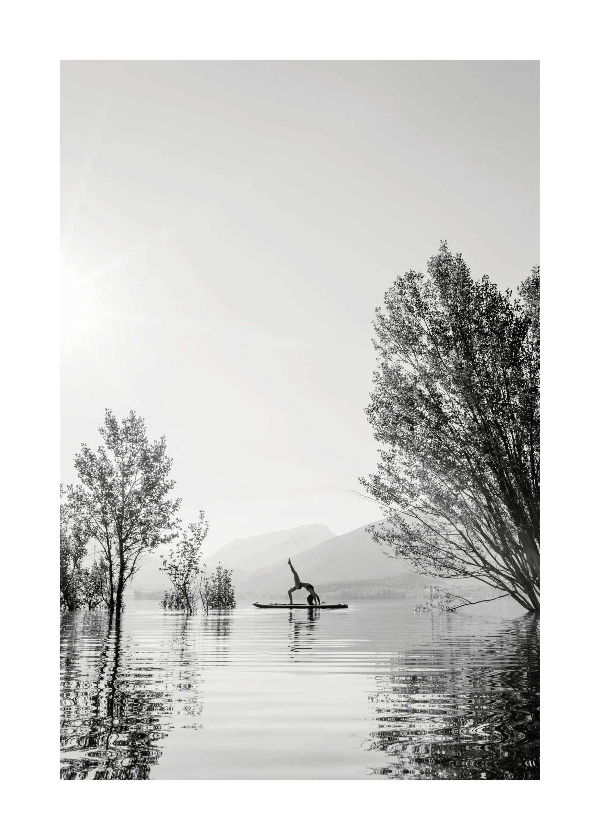 Black and white photograph of a woman doing a yoga pose on a surfboard in a lake, surrounded by trees and mountains.