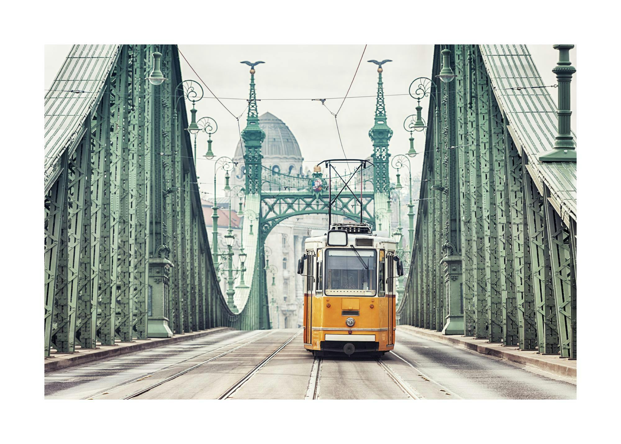 Valokuva keltaisesta vintage-raitiovaunusta vihreällä sillalla Budapestissa, kaupunkimaisema taustalla.